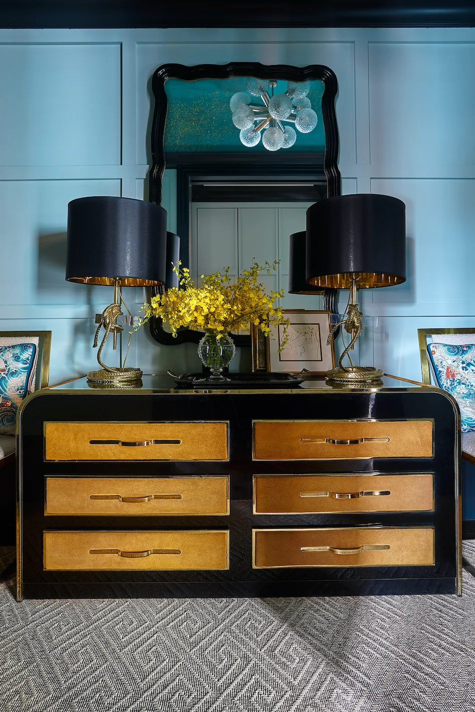 A high-gloss black and gold console table with two black-shaded lamps stands against a light blue paneled wall beneath a curved black mirror.