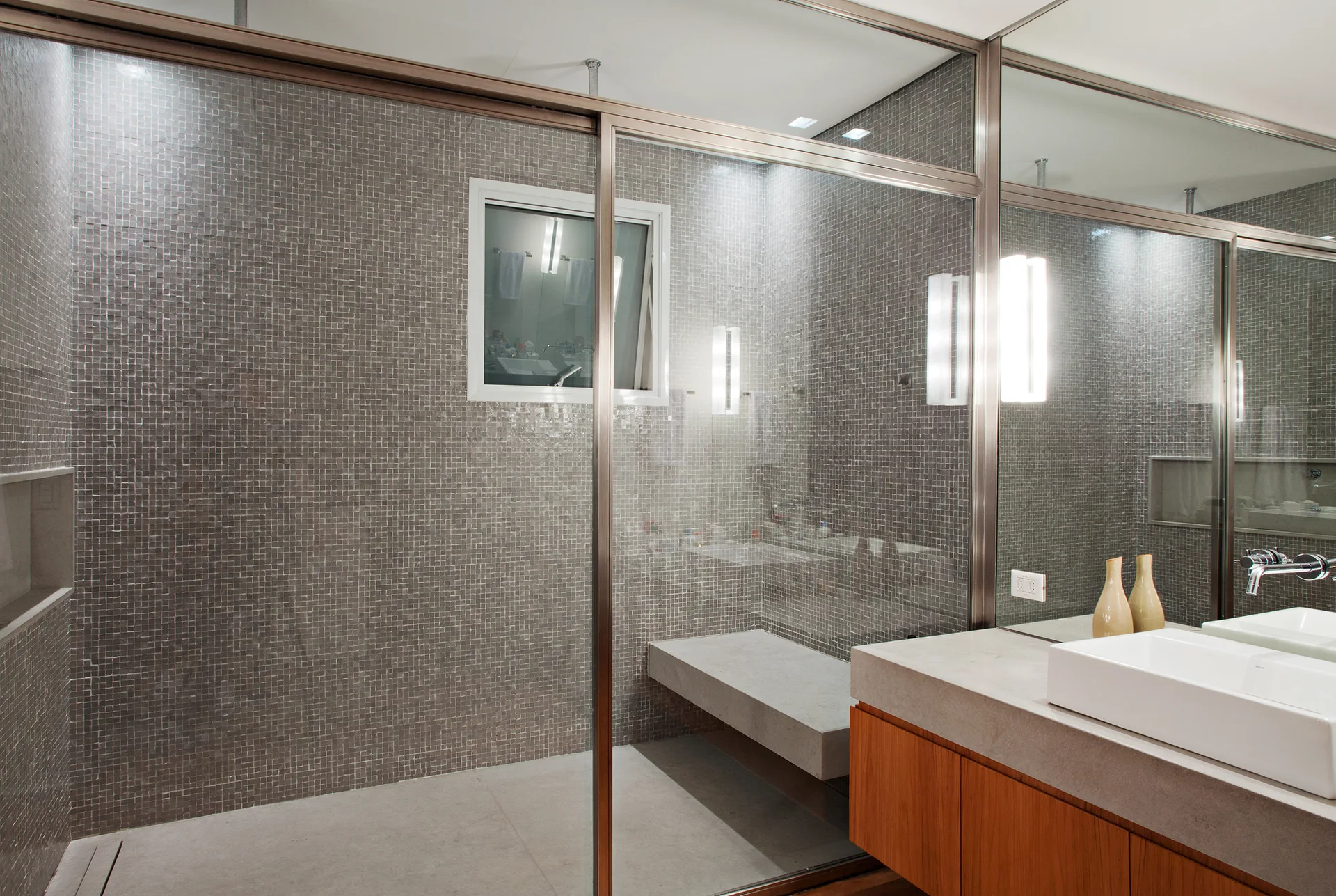 A contemporary bathroom featuring a glass shower enclosure with grey mosaic tile, a floating concrete bench, and a wooden vanity with a white vessel sink.