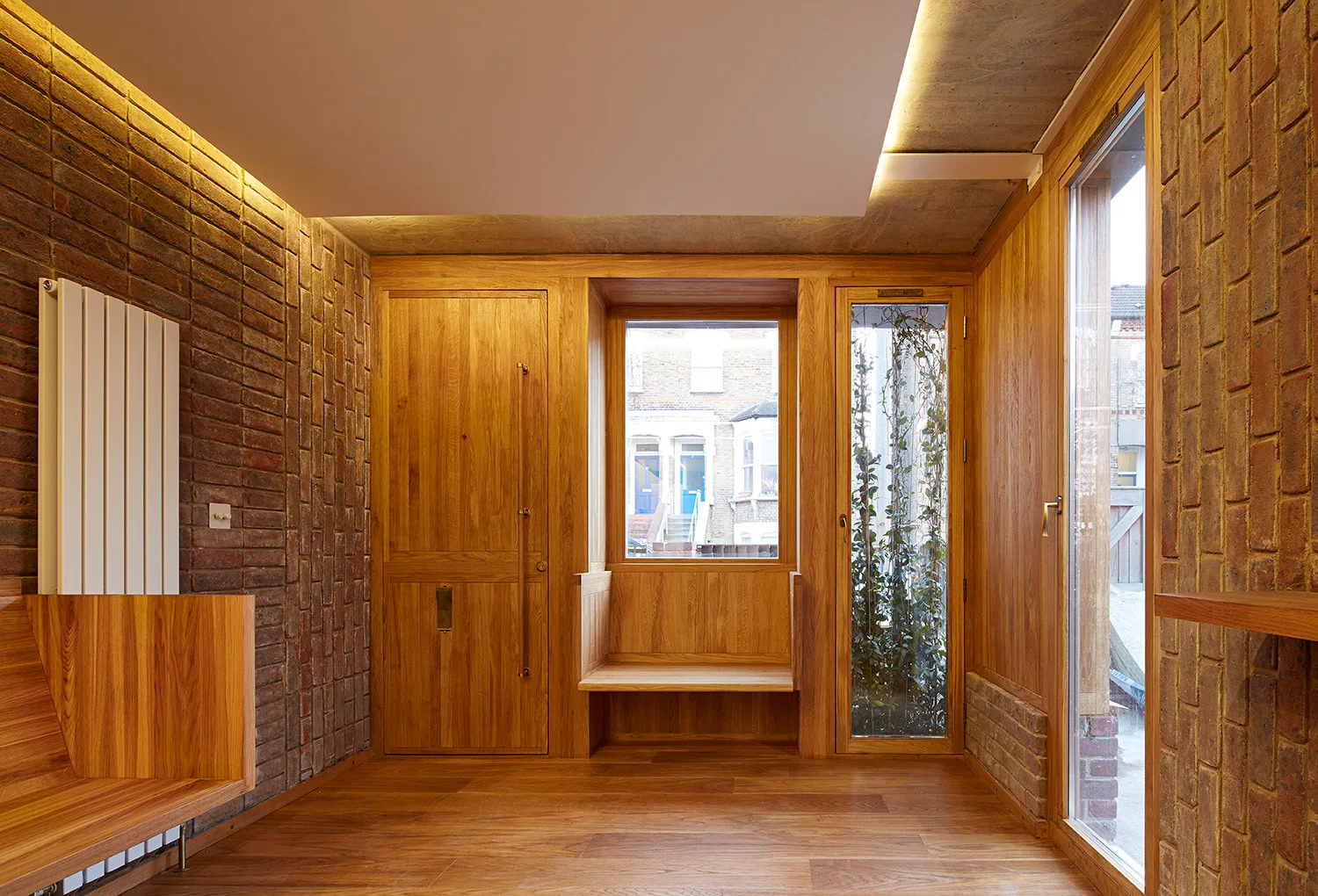 A modern entryway features natural wood-paneled walls, an integrated window bench, exposed brick walls, and indirect cove lighting.