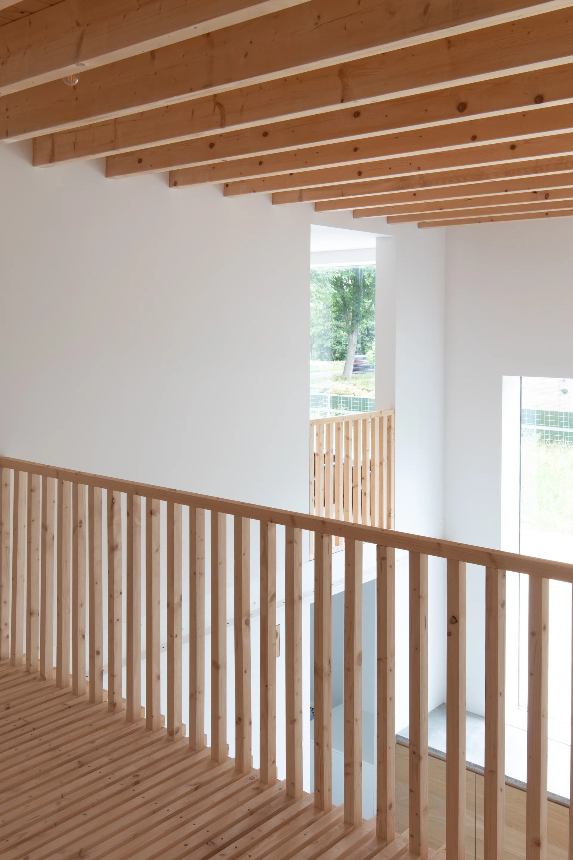 A minimalist wooden landing features a slatted floor, vertical baluster railing, and exposed ceiling beams against bright white walls.