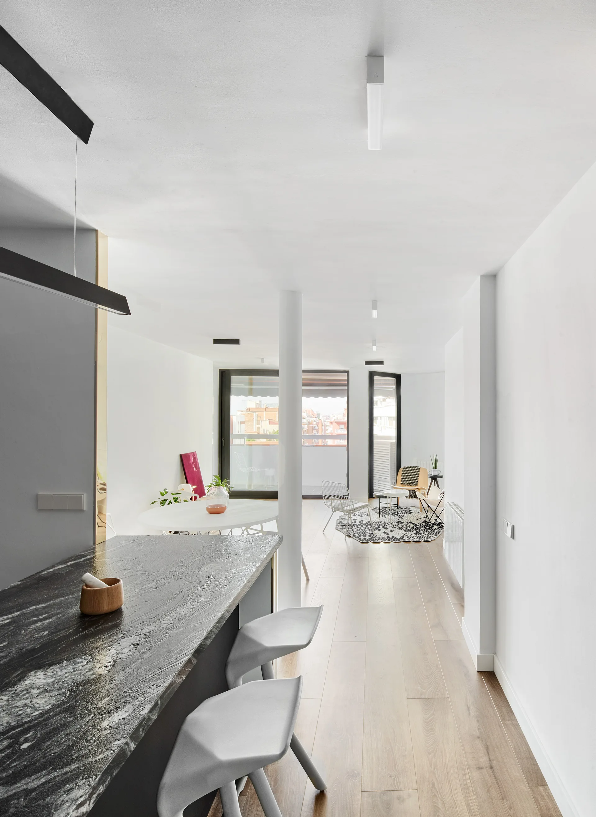 A dark stone kitchen island with grey stools leads into a bright, open-plan space with a round dining table, white column, and a distant seating area.