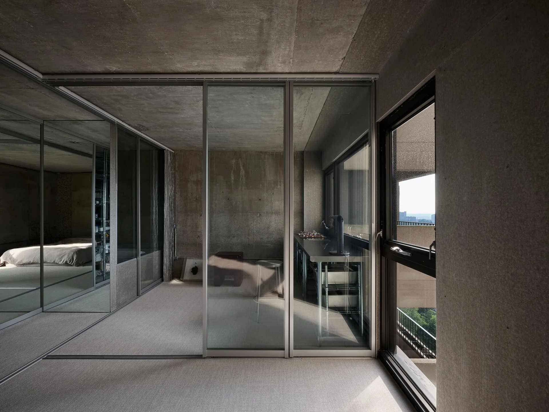 A minimalist room featuring exposed concrete walls and ceiling, a large sliding glass partition, and a mirrored wall reflecting a bed.
