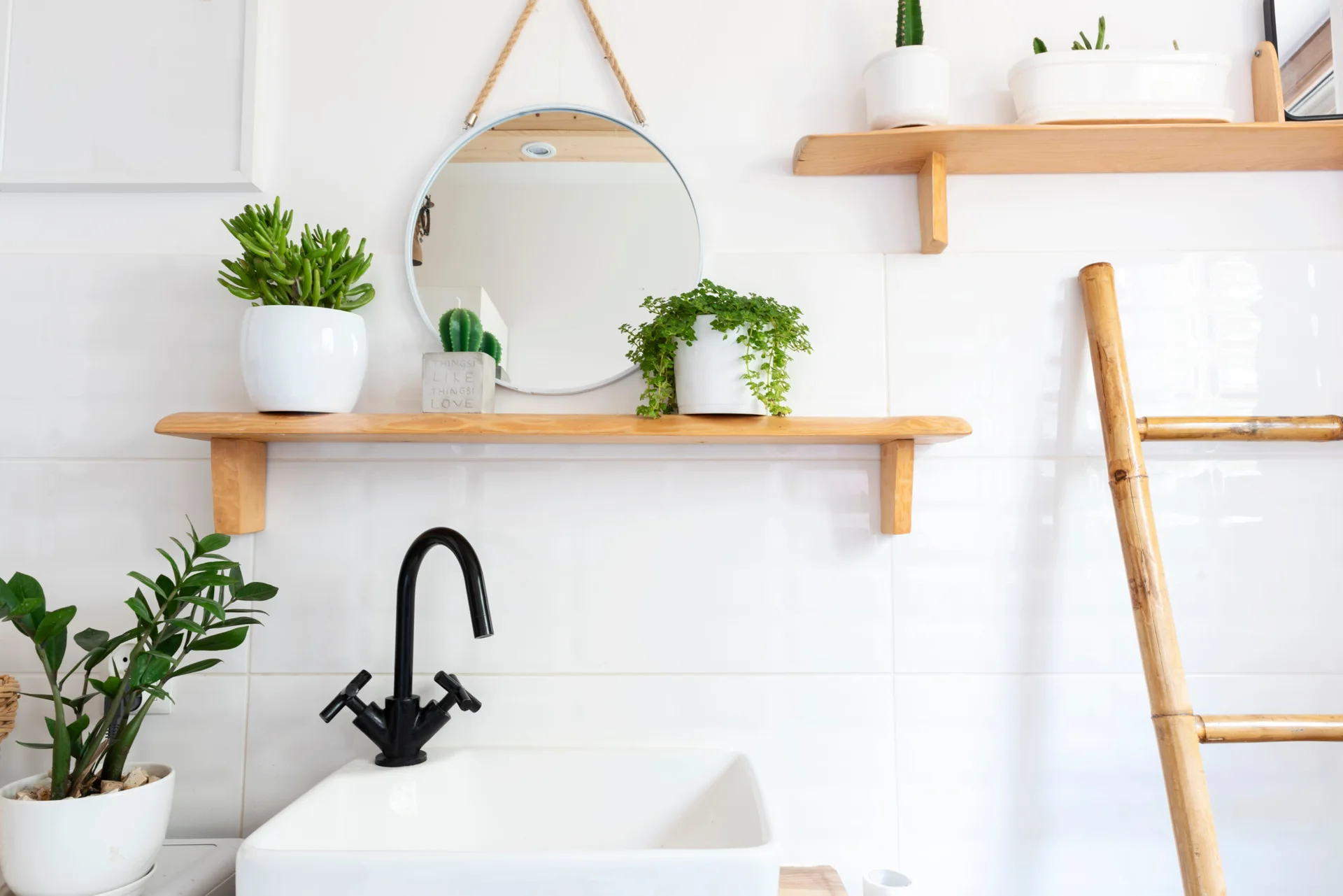 A white ceramic bathroom sink with a black faucet sits below wooden shelves, a round rope-hung mirror, and potted green plants against a tiled wall.