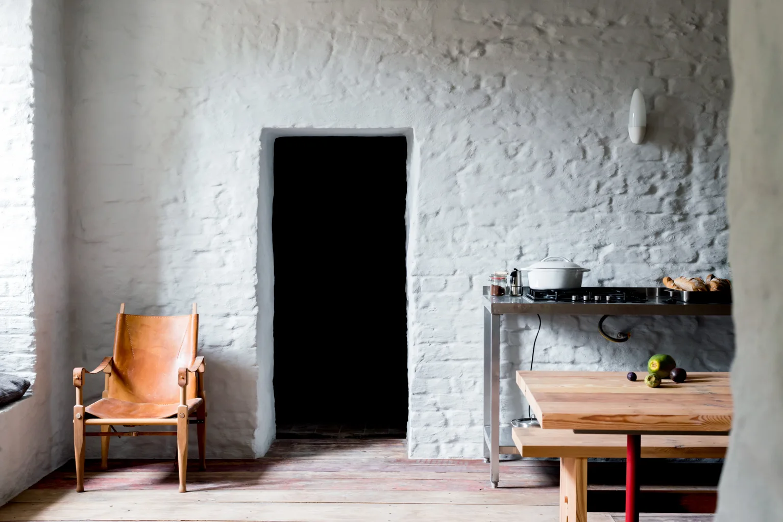 A rustic dining space features a textured white brick wall with a dark doorway, a leather sling chair, and a wooden table.