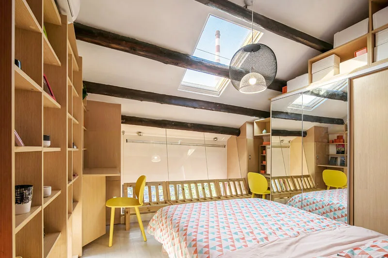 A loft bedroom with a sloped ceiling features exposed wood beams, skylights, built-in wooden shelves, and a mirrored wardrobe reflecting a yellow chair.