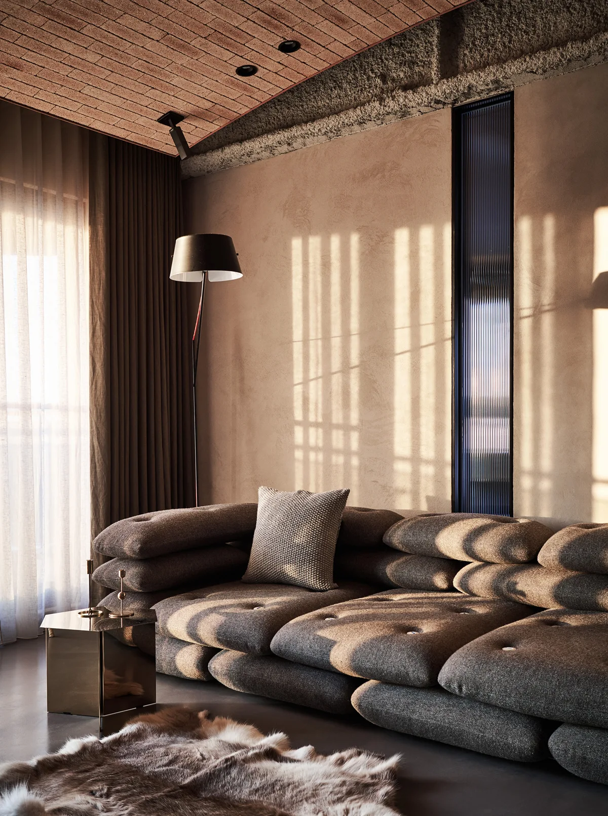A low-profile brown cushion sofa sits against a textured plaster wall next to a slender vertical window and a metallic side table.