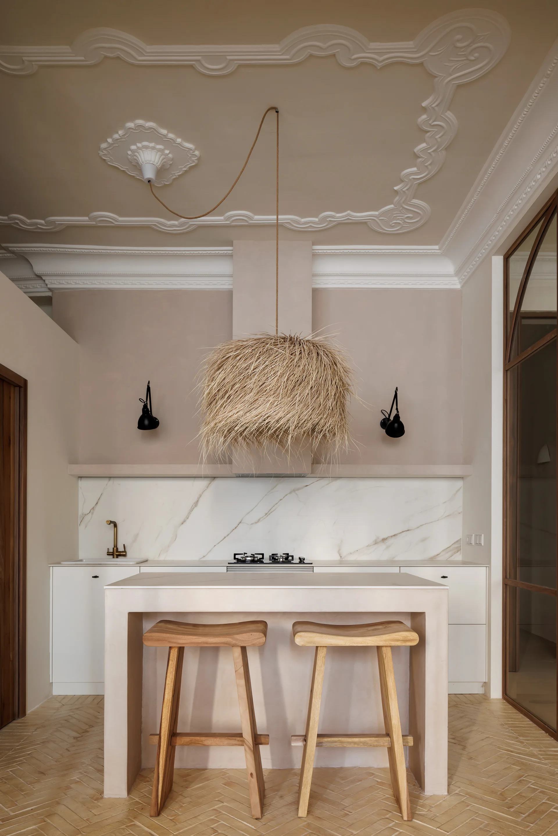 A beige kitchen features a central island with wooden stools, a large textured straw pendant light, and decorative white plaster ceiling molding.