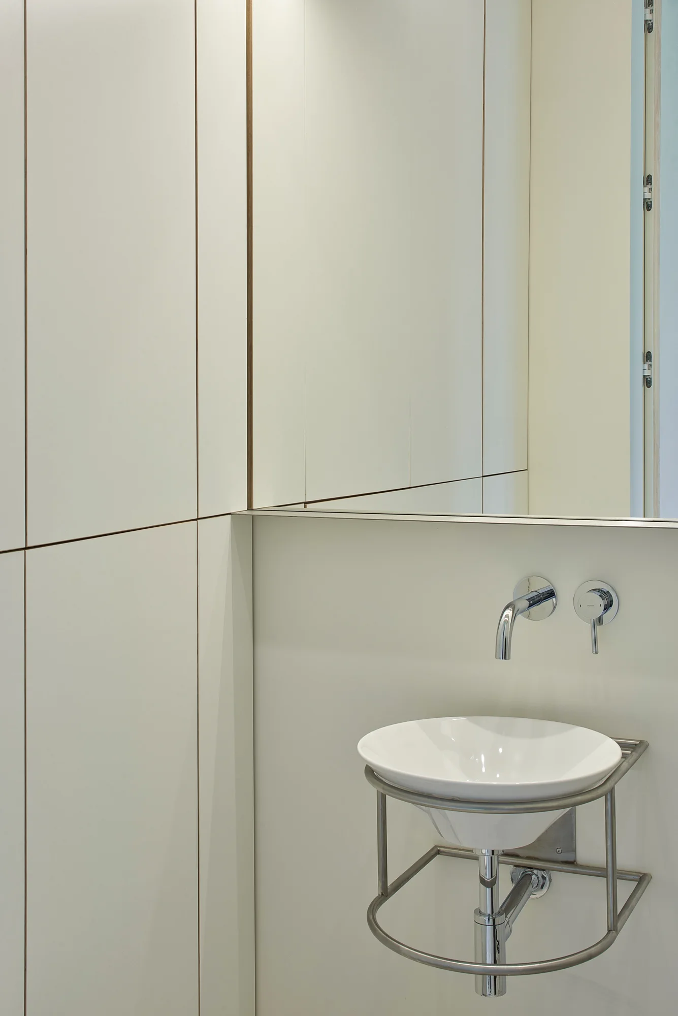 A white ceramic vessel sink on a metal frame is set against cream-colored paneled walls with a wall-mounted chrome faucet and integrated mirror.