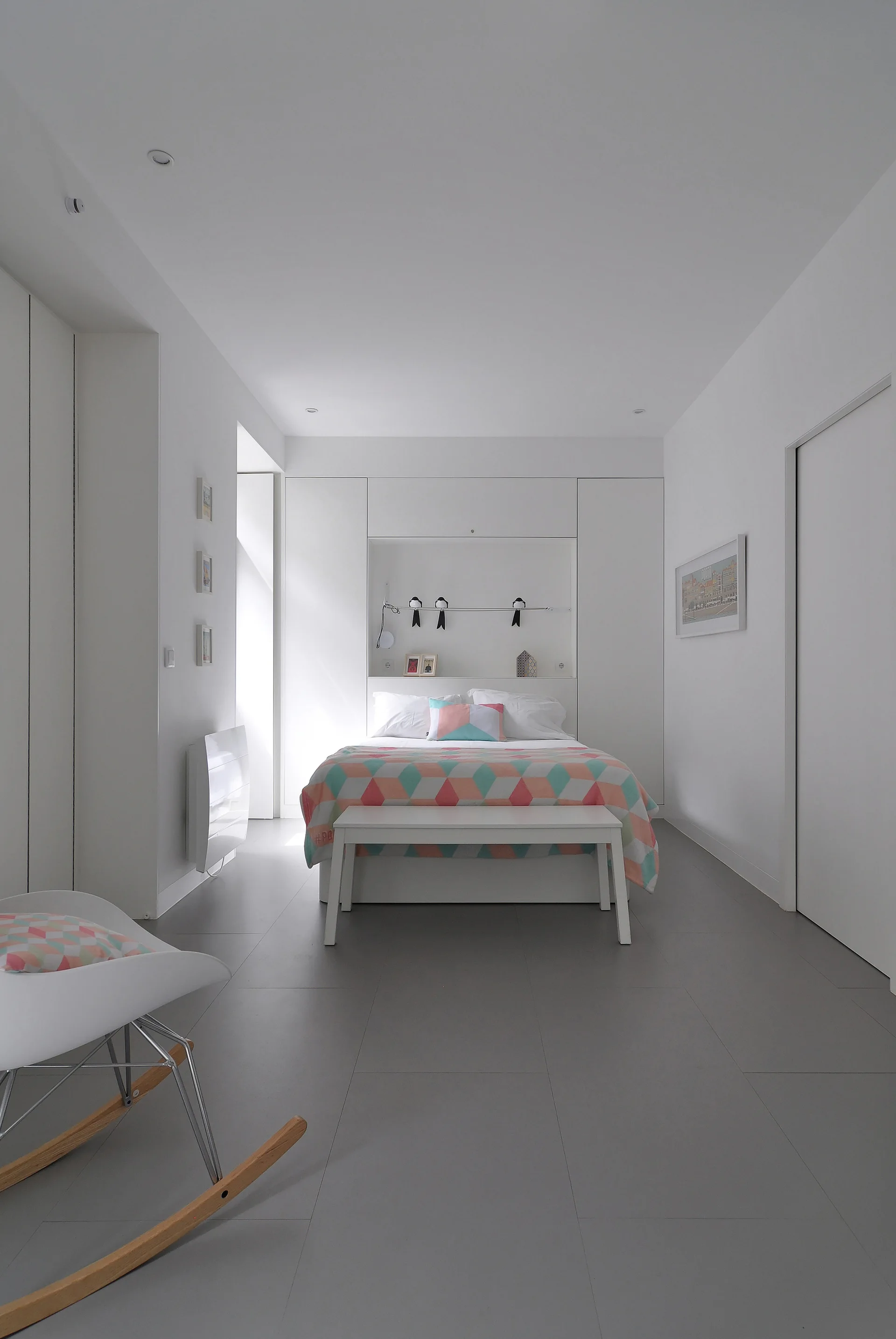 A minimalist white bedroom features a bed recessed into a built-in wardrobe niche, a white bench, and a rocking chair on a grey tiled floor.