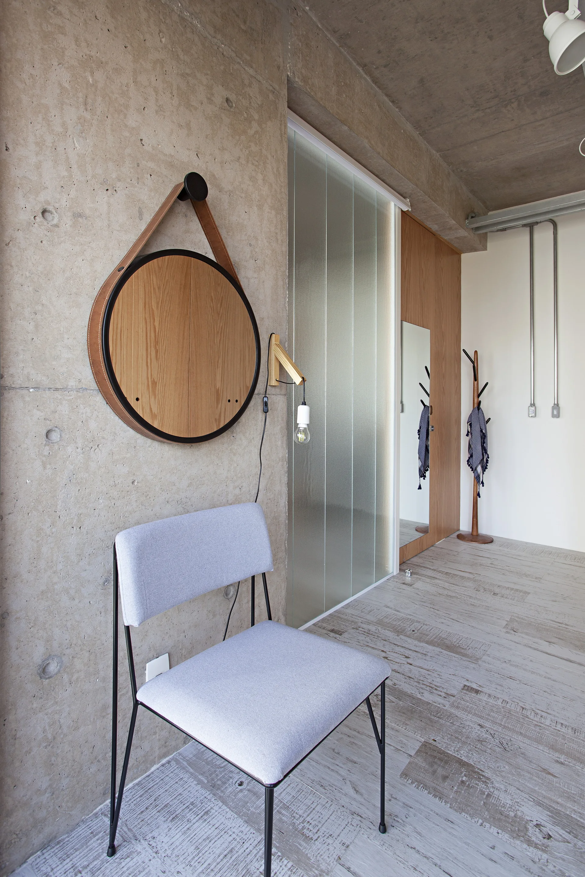 A minimalist entryway features a textured raw concrete wall with a round wood-framed mirror, a grey upholstered chair, and a frosted glass sliding door.