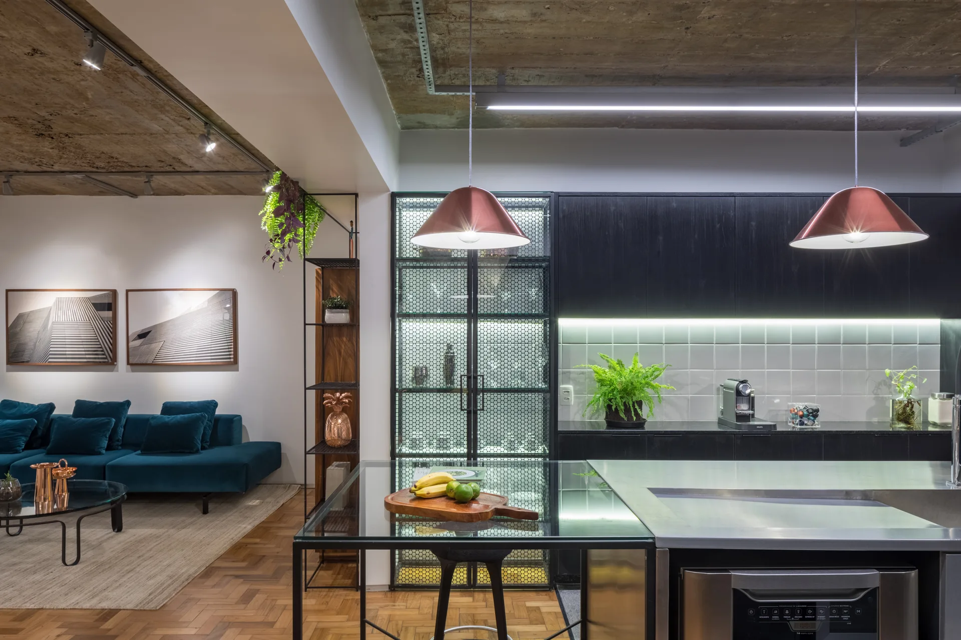 An industrial kitchen features a stainless steel island, black cabinetry, copper pendant lights, and an exposed concrete ceiling looking into a living room.