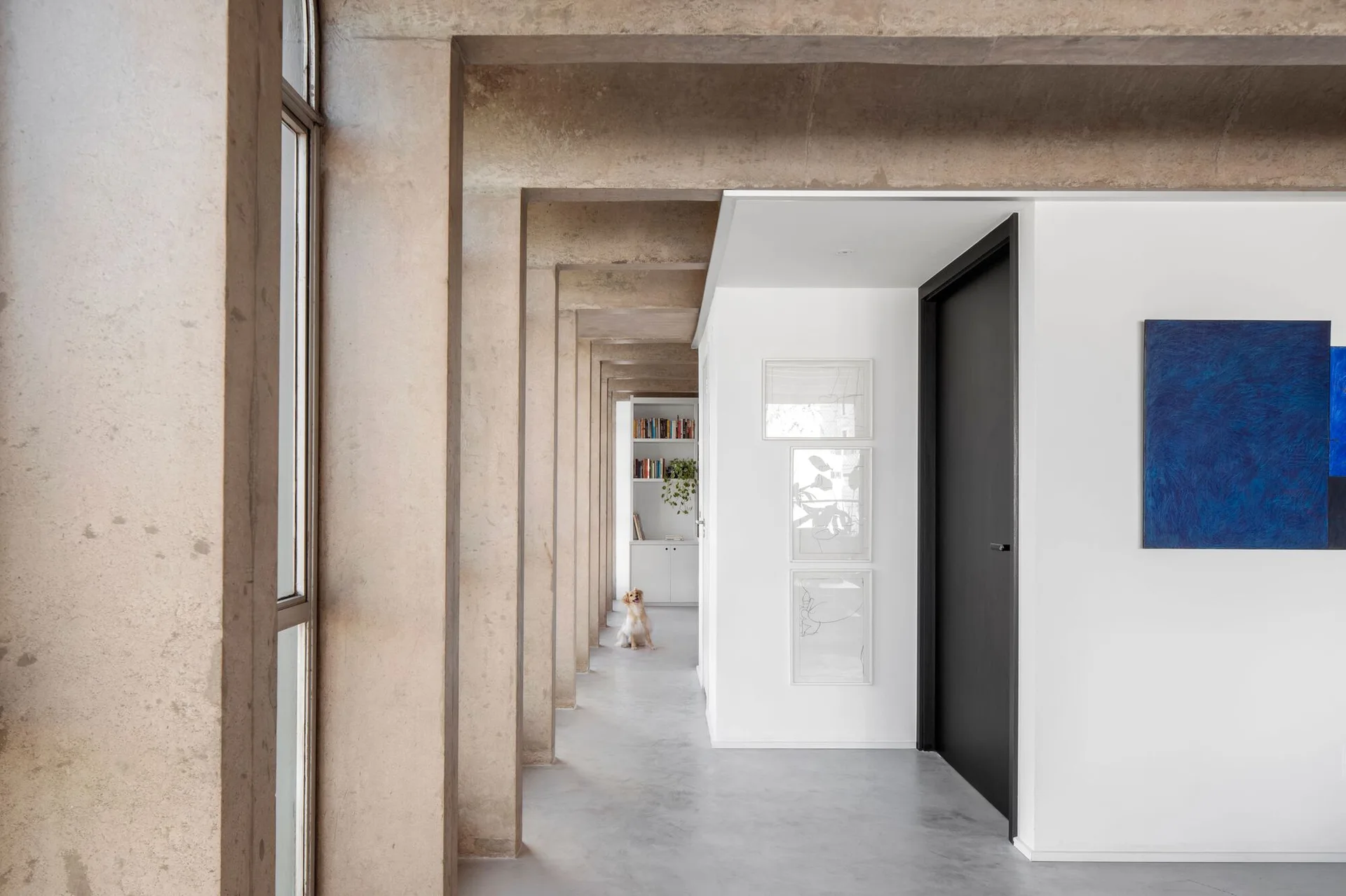 A minimalist hallway featuring structural concrete columns, smooth white walls, a matte black door, and a vibrant blue painting.