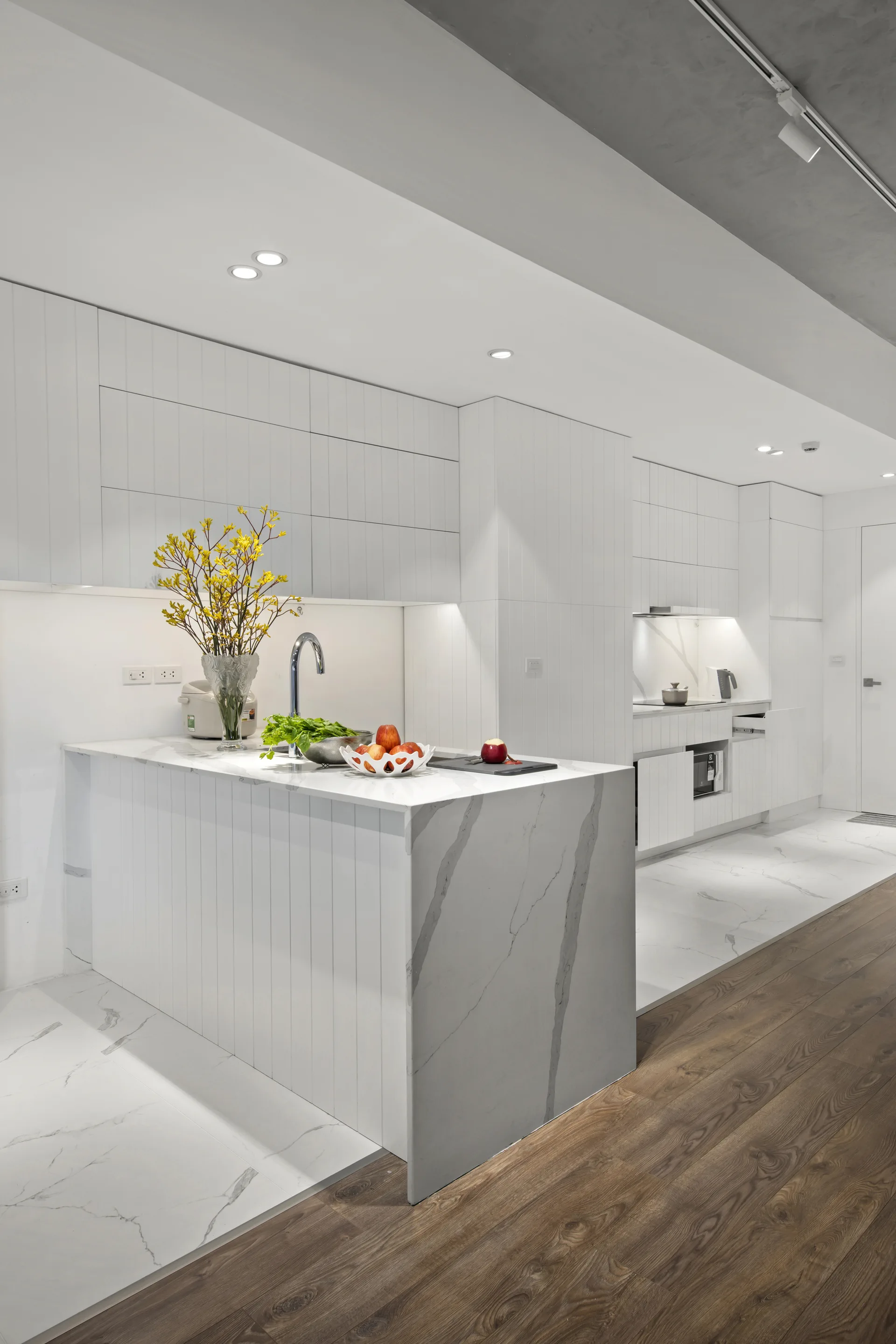 A modern white kitchen featuring a marble waterfall island, vertical slat cabinetry, and a floor transition from marble tile to dark wood.