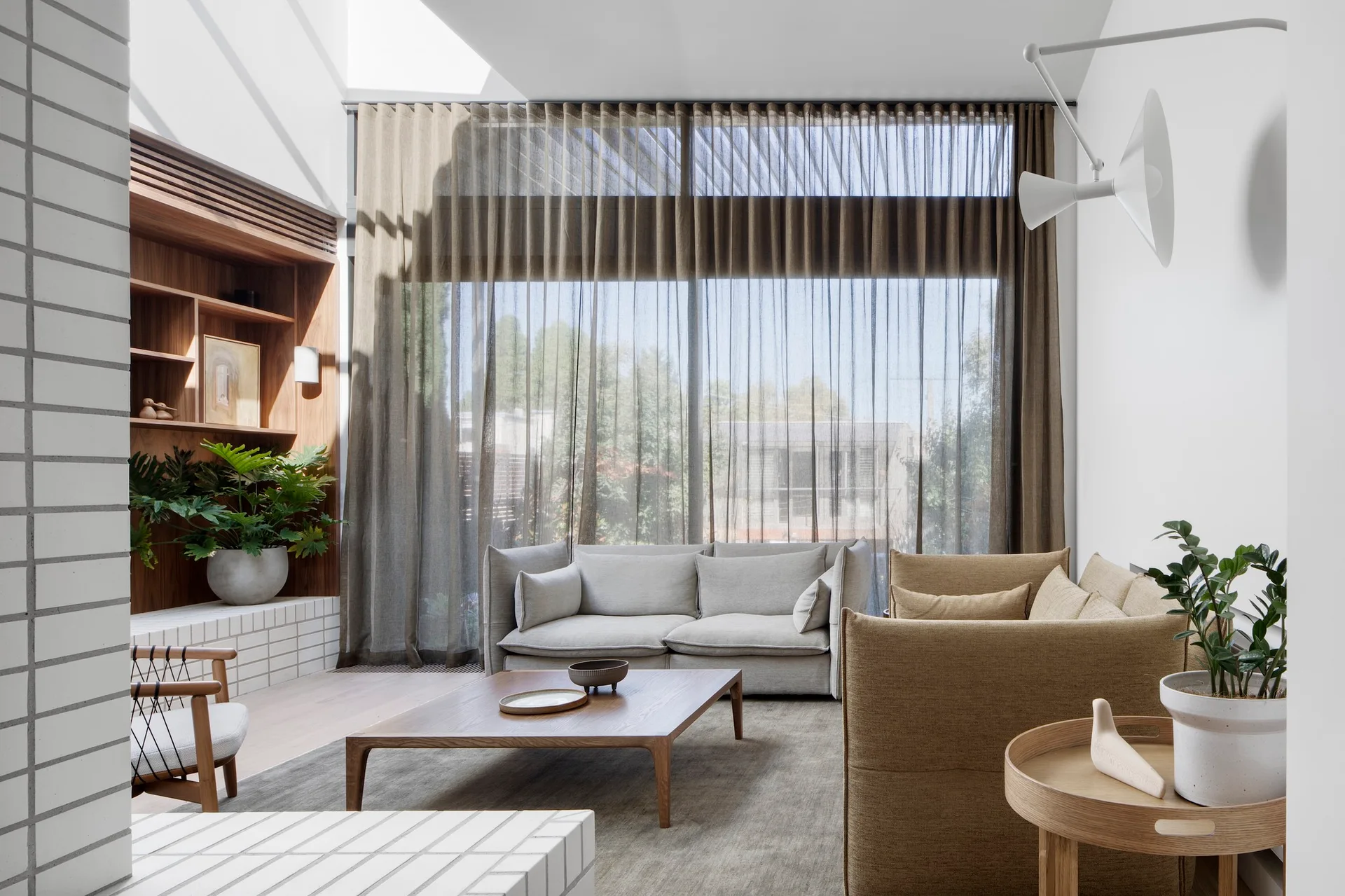 A contemporary living room featuring grey and beige sofas, a low wood coffee table, sheer brown curtains, and a recessed wooden shelving unit.