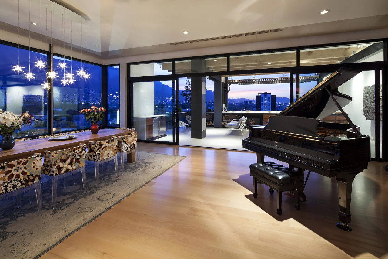 A spacious dining area features a black grand piano, a long wooden table with patterned chairs, and sliding glass doors leading to a balcony.