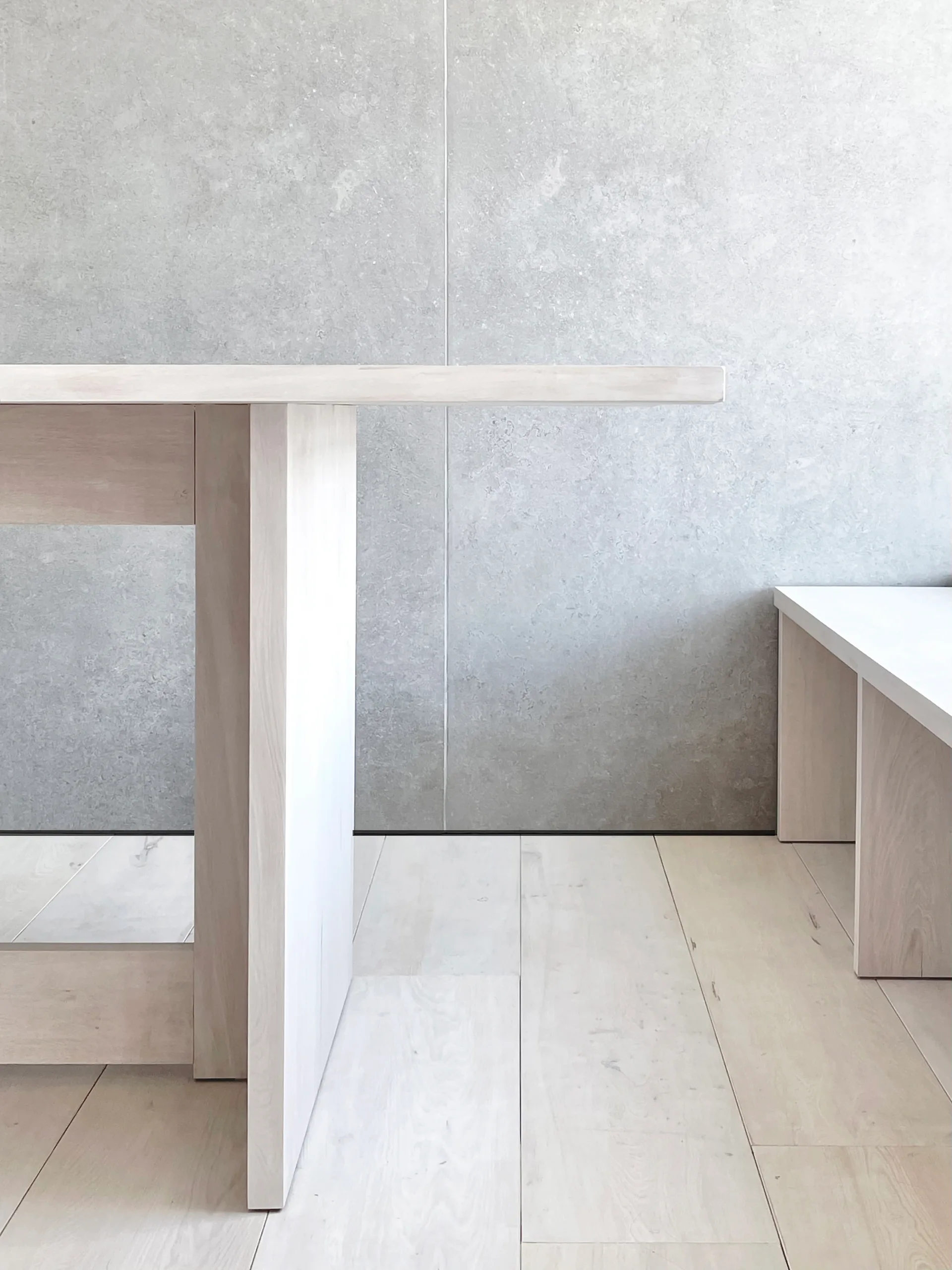 A minimalist light wood dining table and bench set against a large textured grey stone wall panel and matching wood plank flooring.