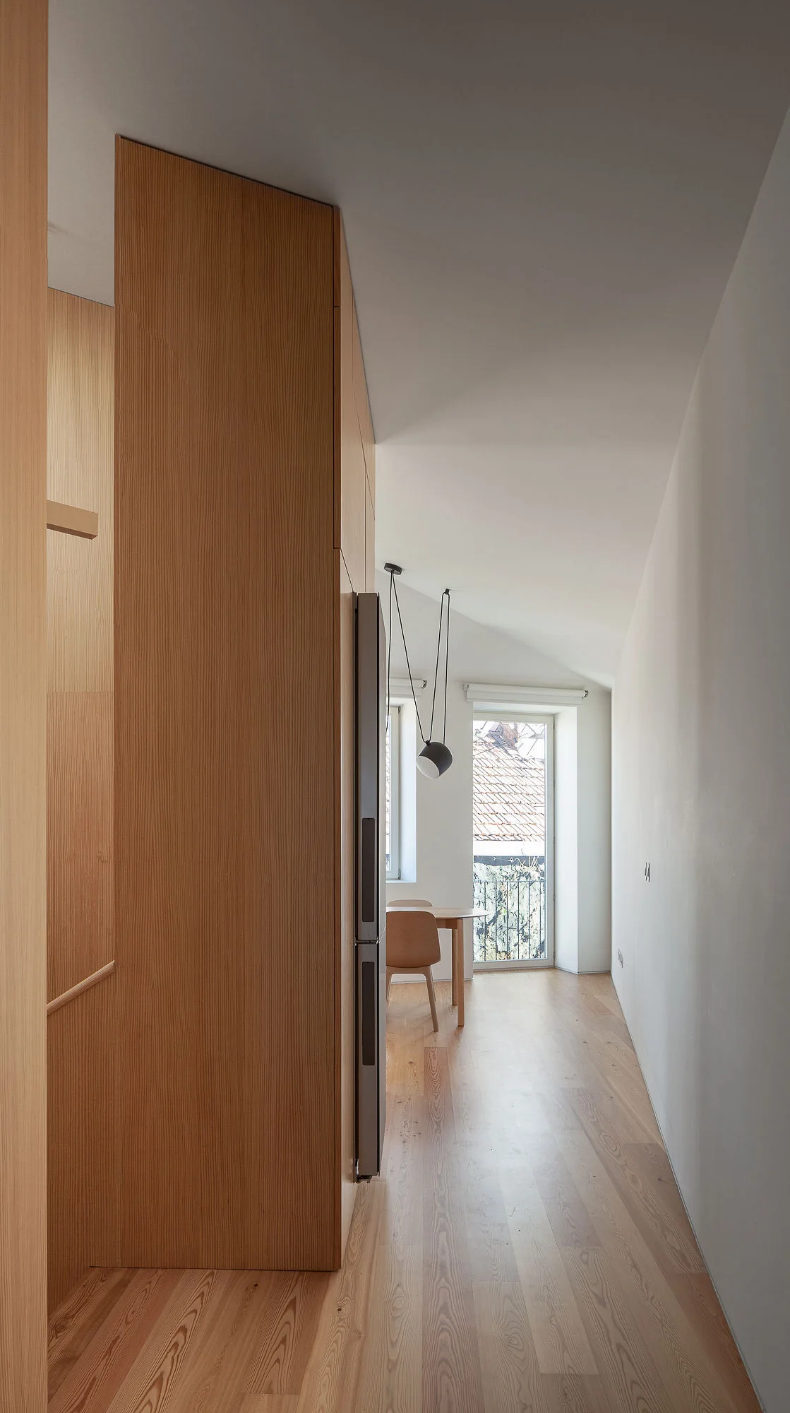 A narrow corridor features floor-to-ceiling wood cabinetry, an integrated refrigerator, and light wood floors leading to a sunlit dining area.