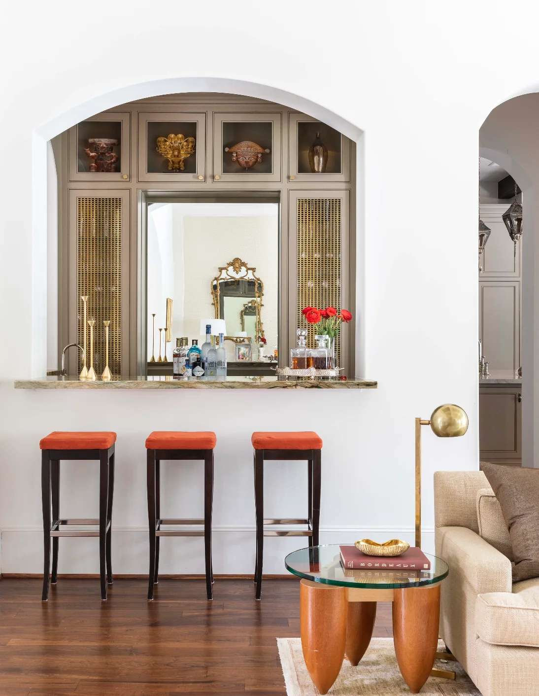 A home bar set inside a white archway features taupe cabinetry with metal grilles, a stone counter, and three dark wood stools with orange cushions.