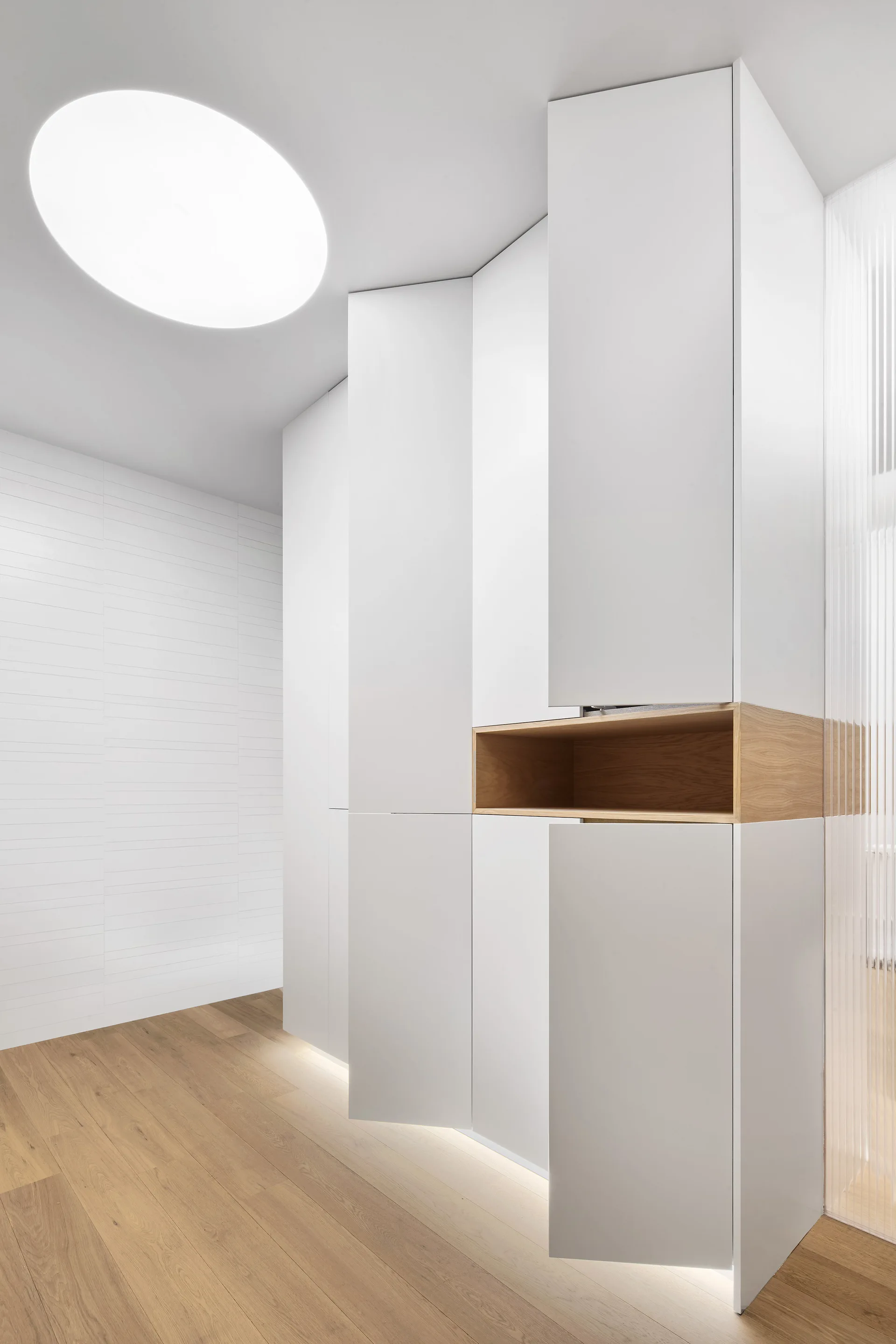 Floor-to-ceiling white cabinetry in a hallway features a recessed light wood niche, toe-kick lighting, and natural wood flooring.