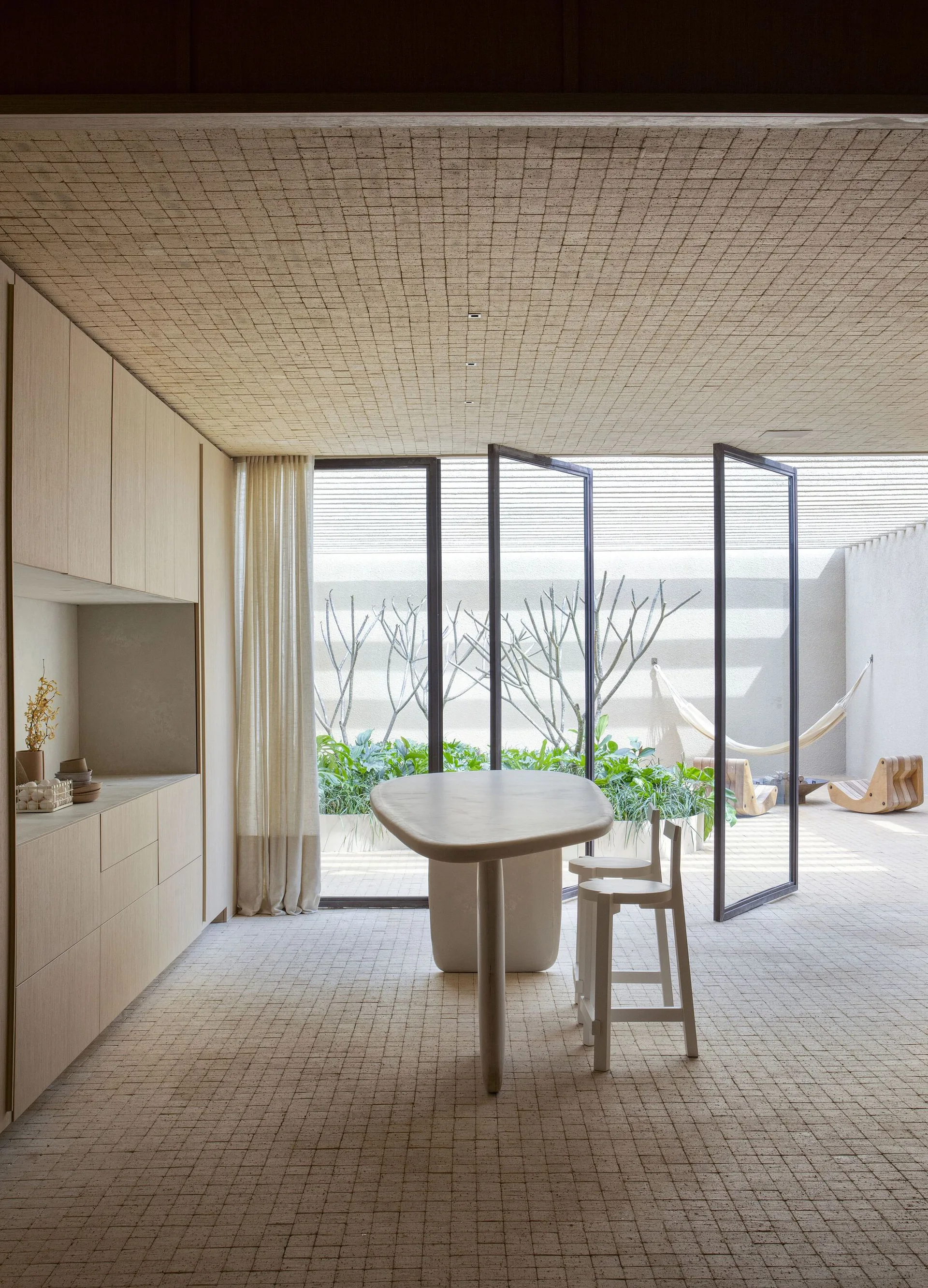 A minimalist kitchen features matching stone-tiled floors and ceilings, large pivoting glass doors, and a sculptural organic dining table.