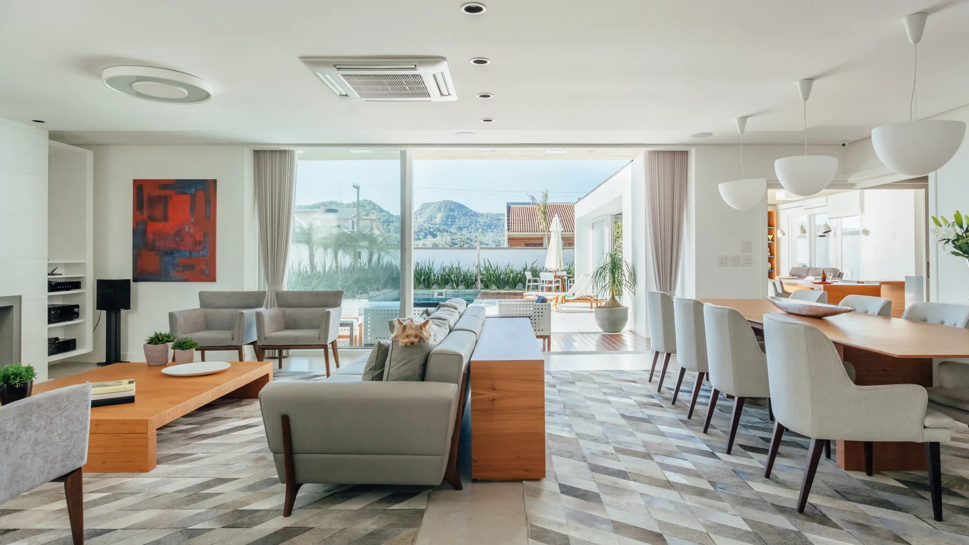An open-concept living and dining area features a grey sofa with a wooden console table, a geometric pattern rug, and a large sliding glass door.