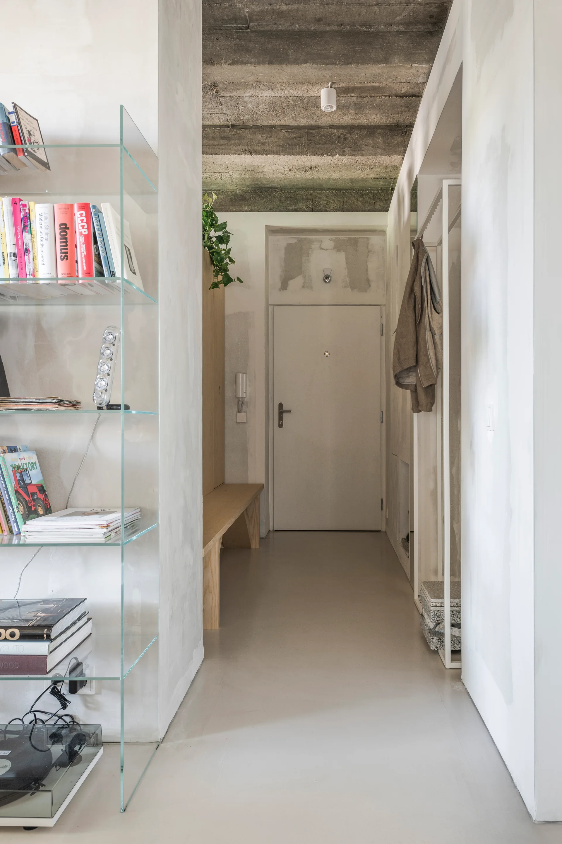 A minimalist residential entry hallway featuring a light wood bench, glass shelving, a white coat rack, and an exposed raw concrete ceiling.