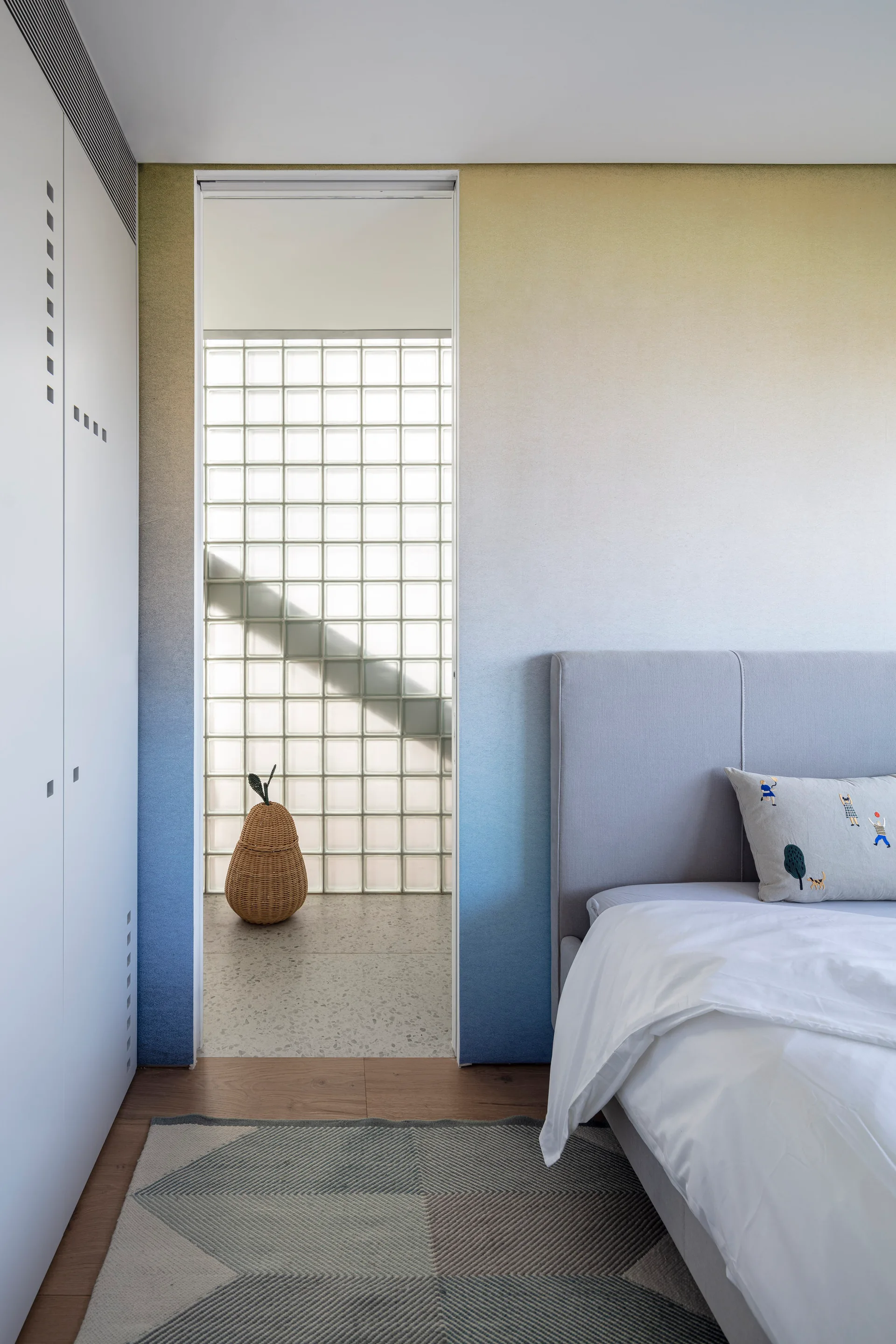 A modern bedroom features a yellow and blue ombre wall, a grey upholstered headboard, and an adjacent room visible through a doorway.
