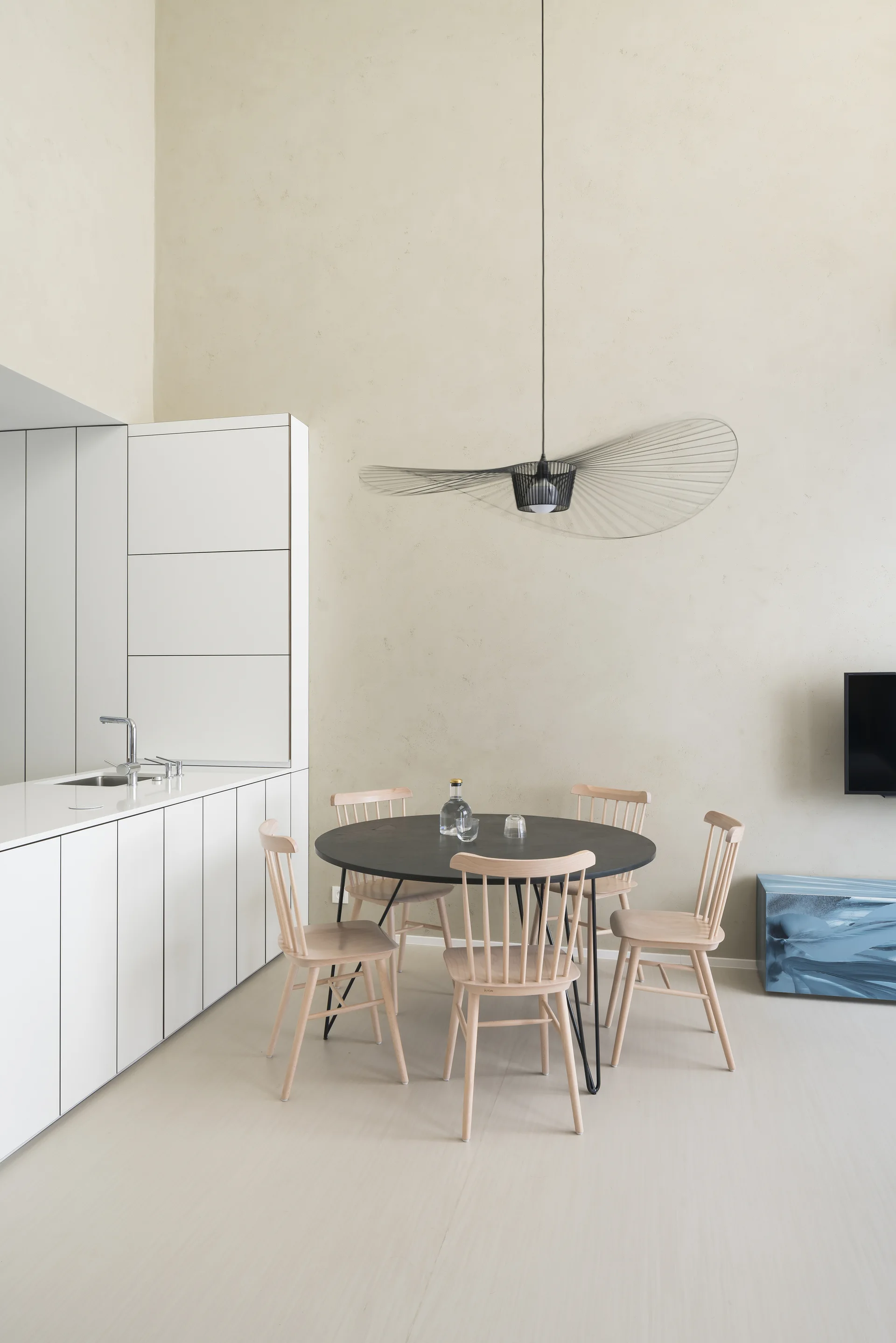 A minimalist dining area features a black round table, light wood spindle chairs, and a large wire pendant light next to seamless white kitchen cabinets.