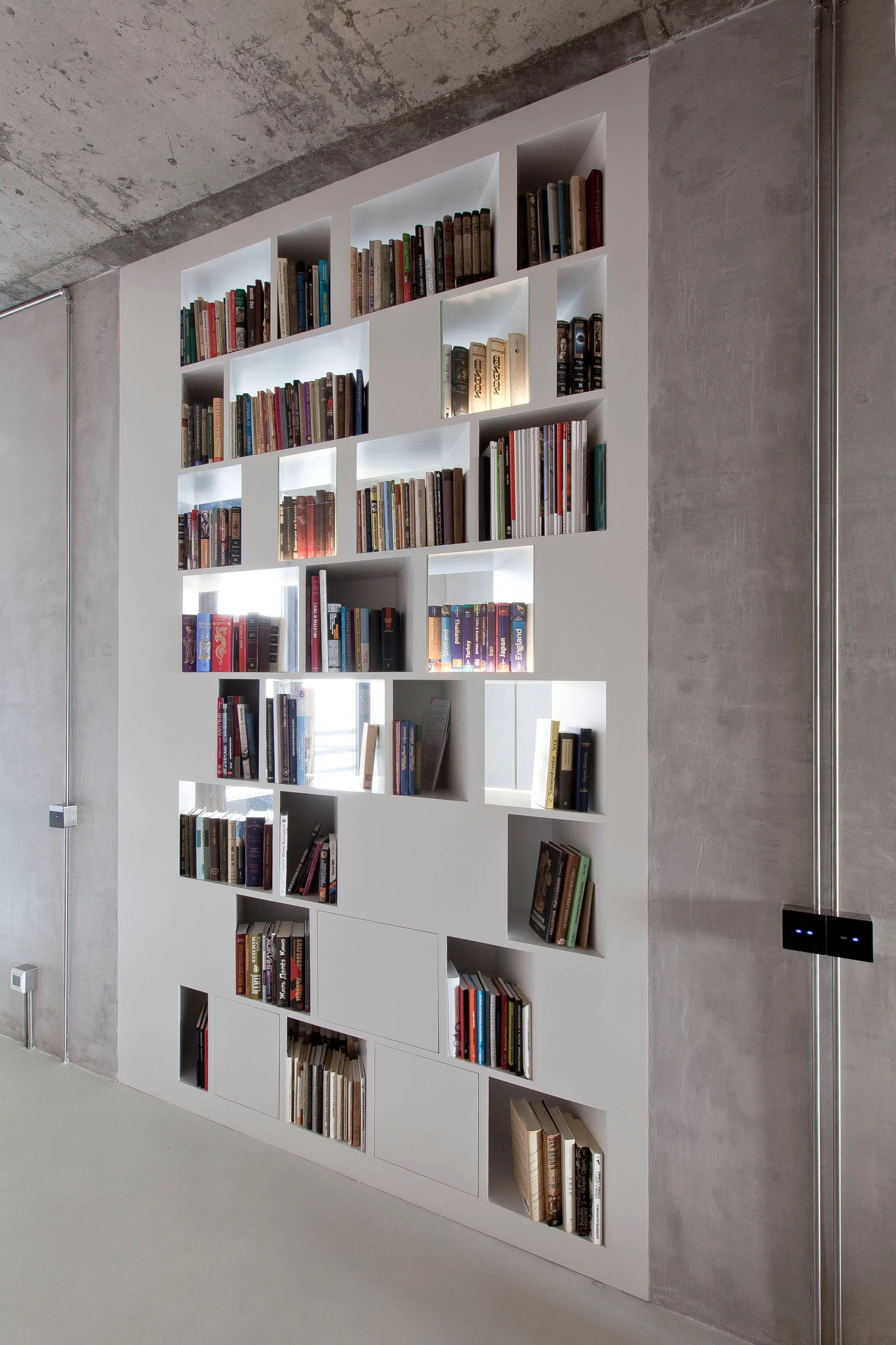 A floor to ceiling white built in bookshelf with asymmetrical compartments and integrated lighting set against a raw concrete wall and ceiling.