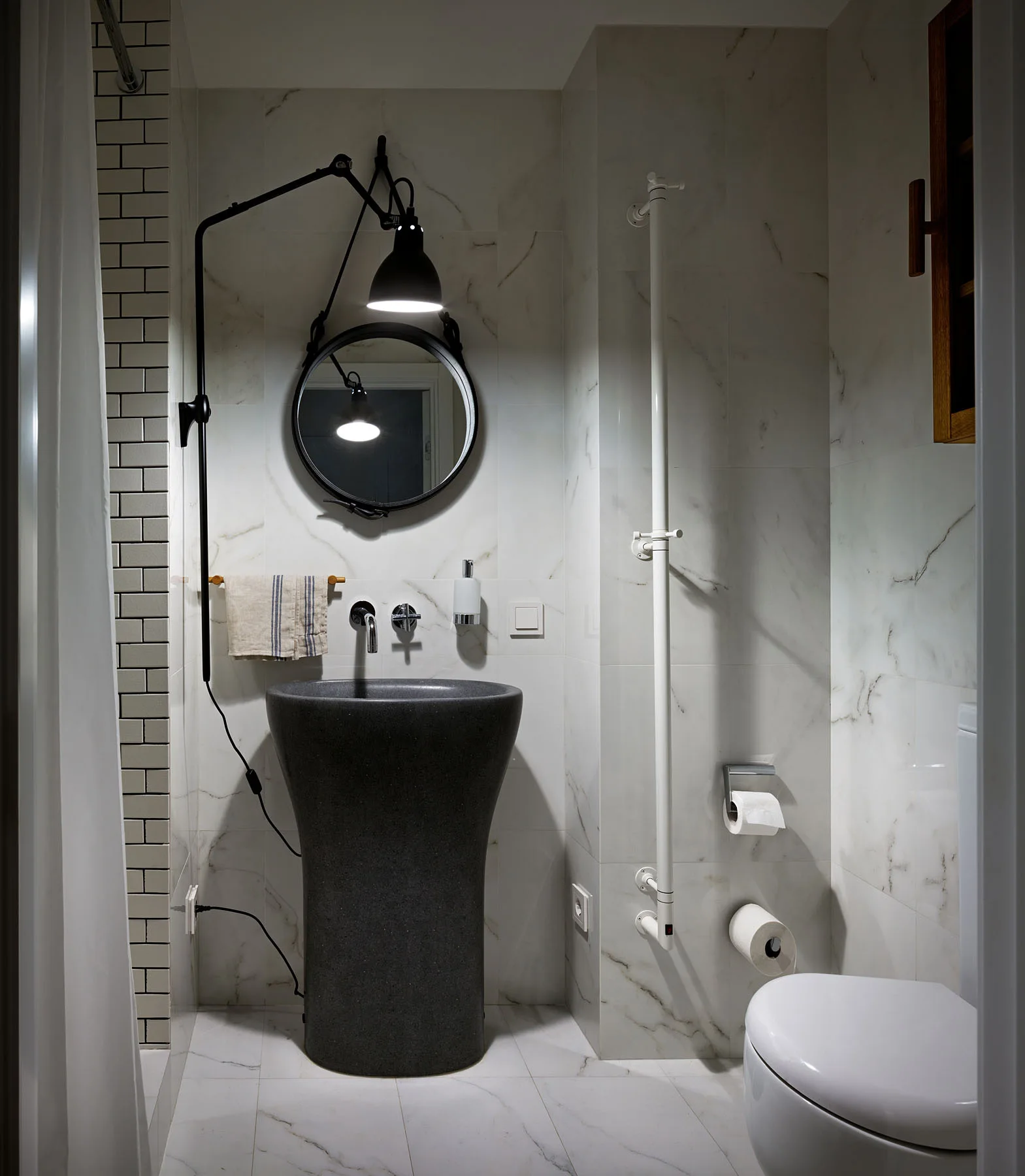 A modern bathroom with white veined marble wall tiles, a sculptural dark gray pedestal sink, a round wall mirror, and a black swing-arm lamp.