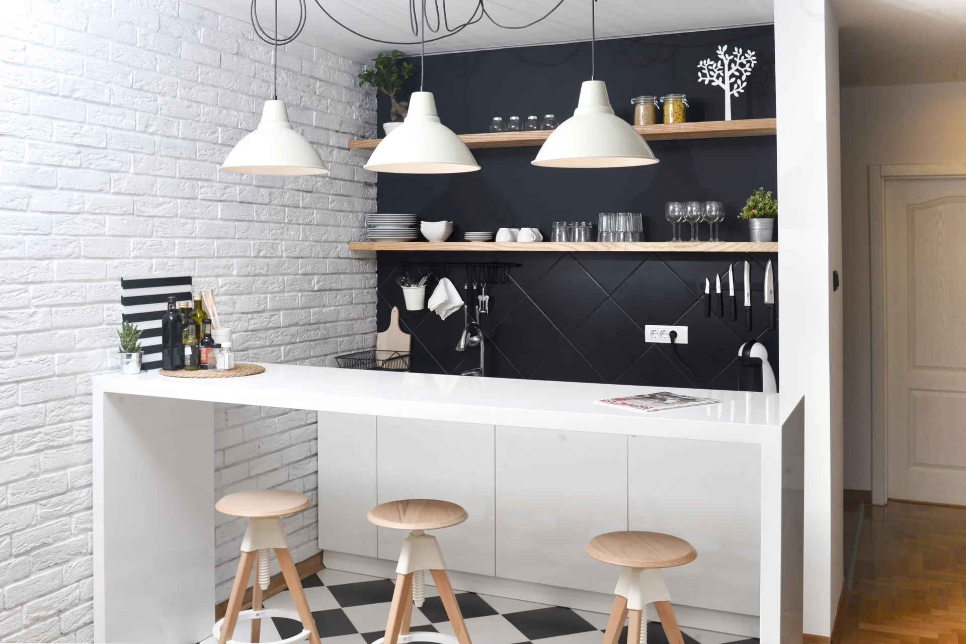 A kitchen breakfast bar features a white counter, wooden bar stools, white brick wall, and a black diamond-patterned tile backsplash.