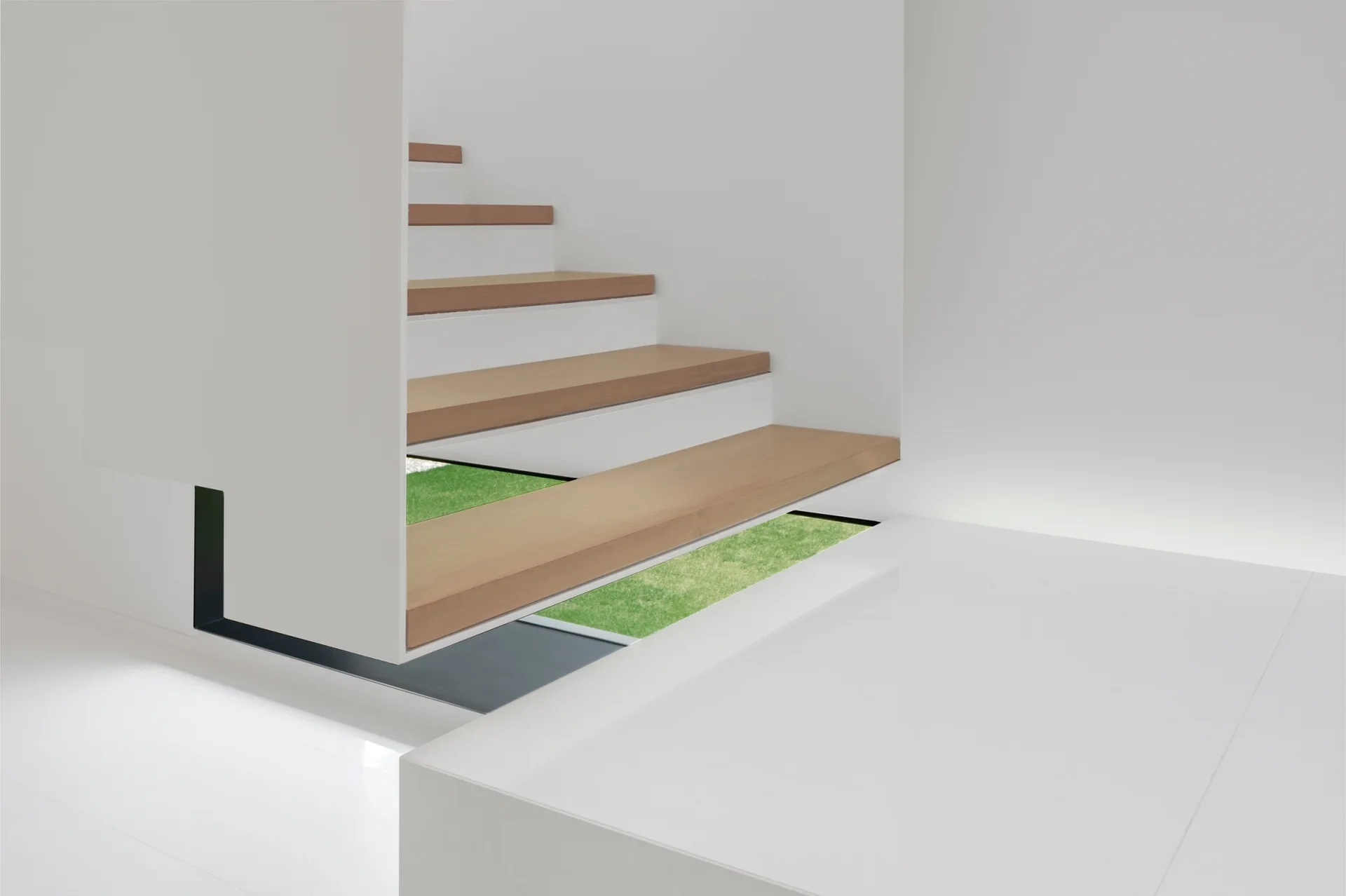 Minimalist staircase with wooden floating treads and white risers, framing a low ground-level window that looks out onto a green lawn.