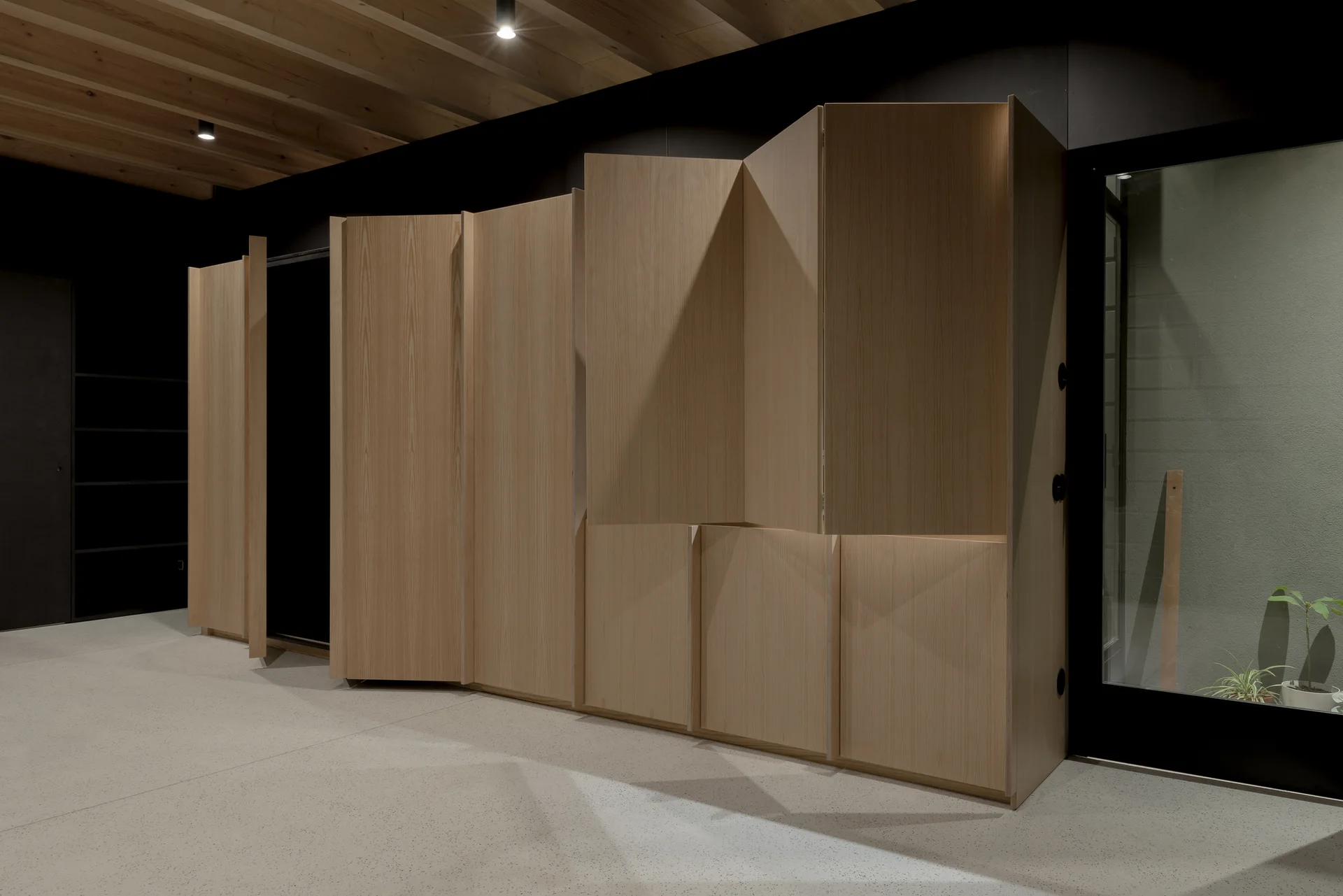 A large faceted light wood cabinet unit stands against a matte black wall under an exposed wood beam ceiling with recessed spotlights.