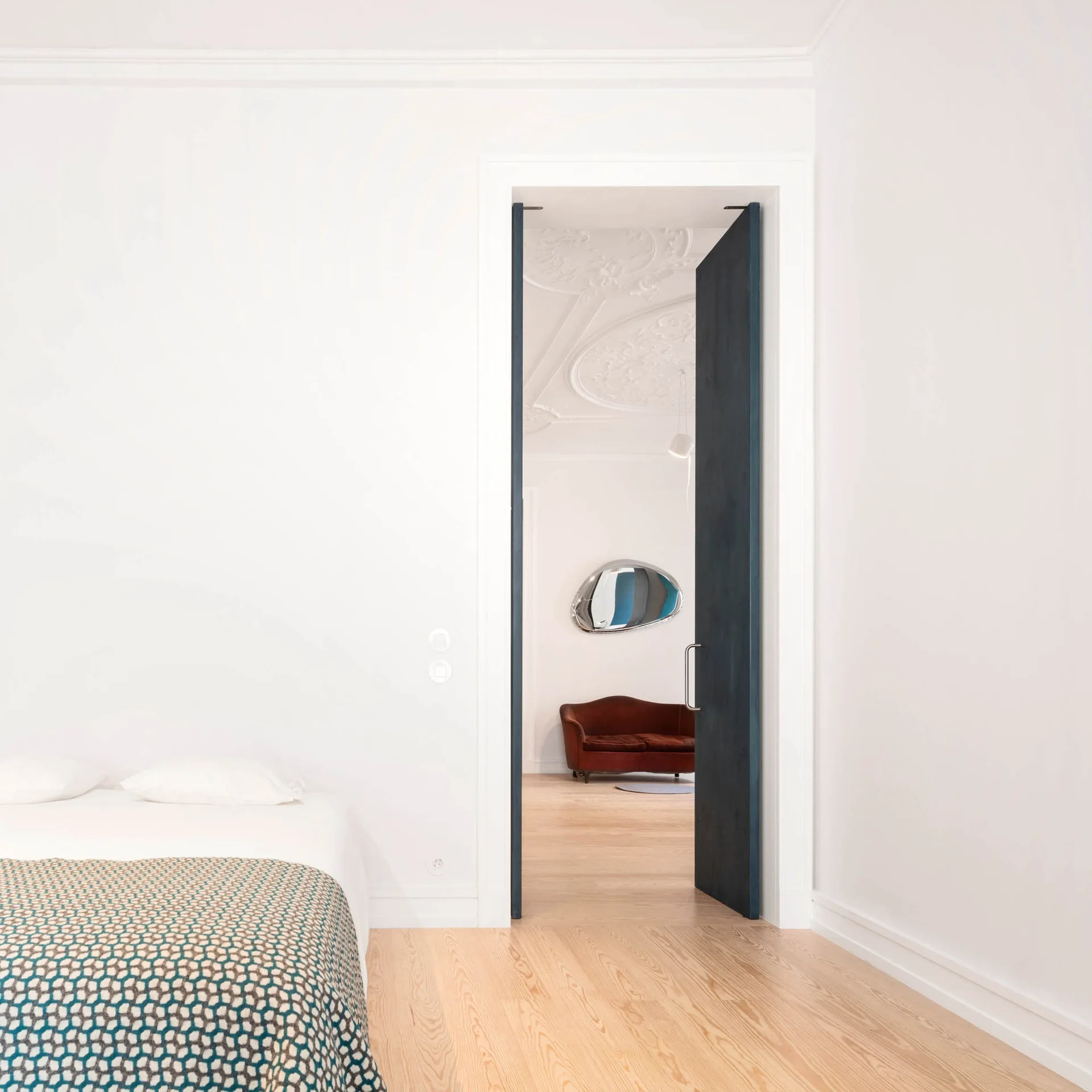 A minimalist white bedroom with light wood flooring features a tall, open dark blue door leading to an adjacent room with a red sofa and ornate ceiling.