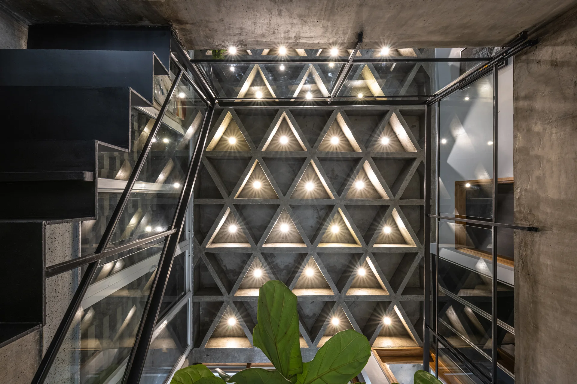 Upward view of a stairwell with a black metal staircase, glass railings, and a concrete ceiling in a triangular geometric pattern with spotlights.