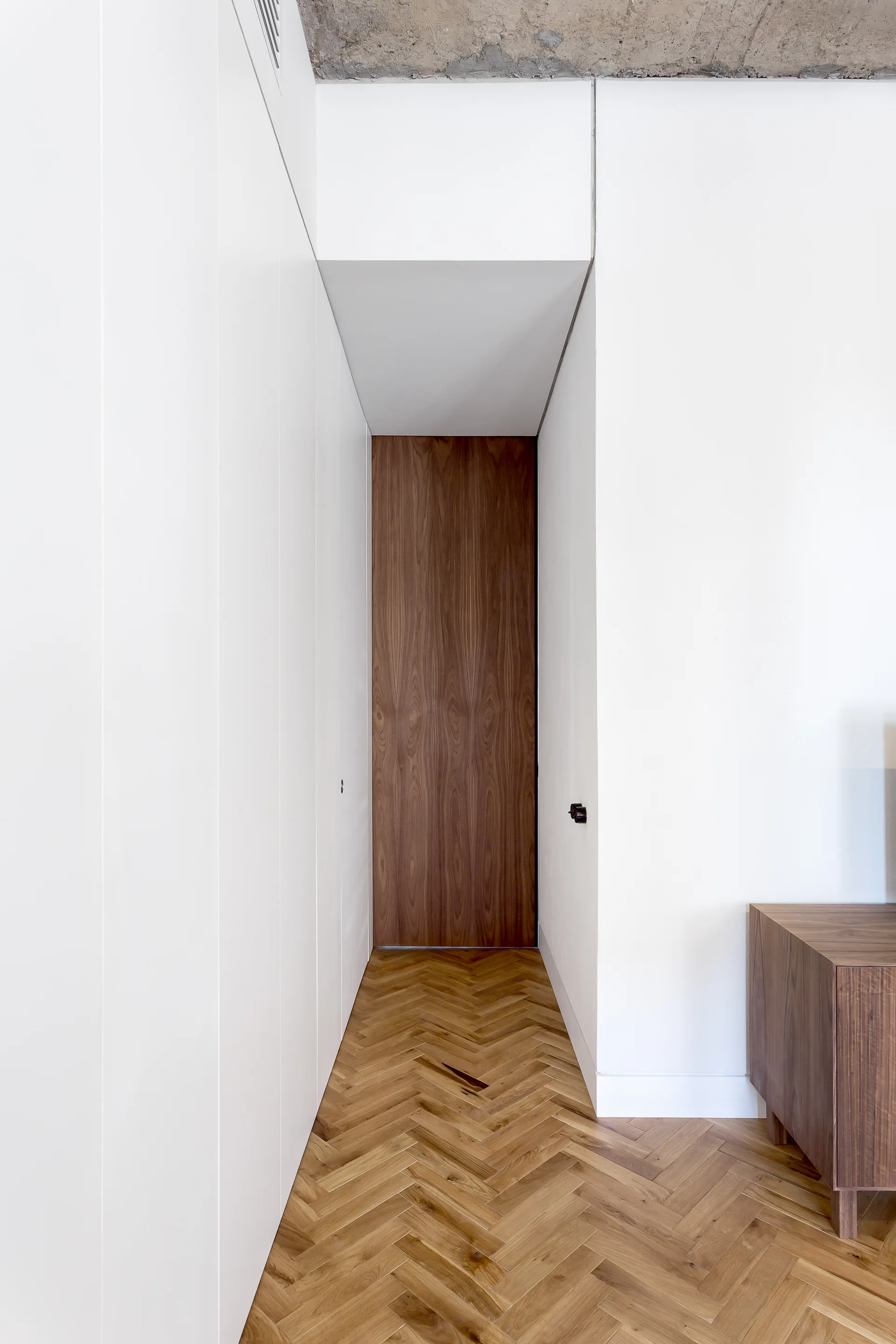 A minimalist white hallway features light oak herringbone parquet flooring, a flush wood grain door at the end, and a raw concrete ceiling above.