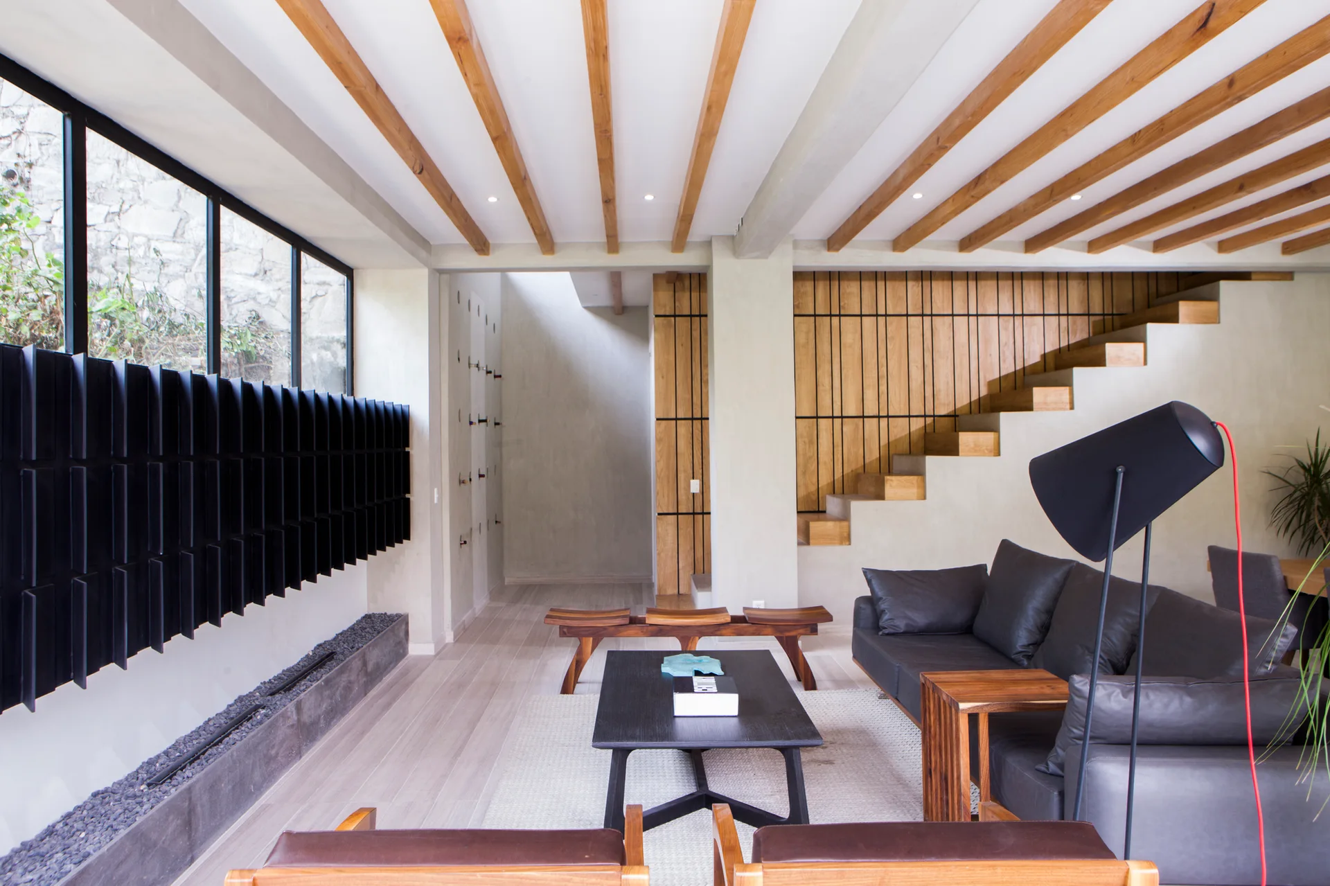 A modern living room features exposed wood ceiling beams, a black leather sofa, and an open riser staircase next to a vertical wood slat wall.