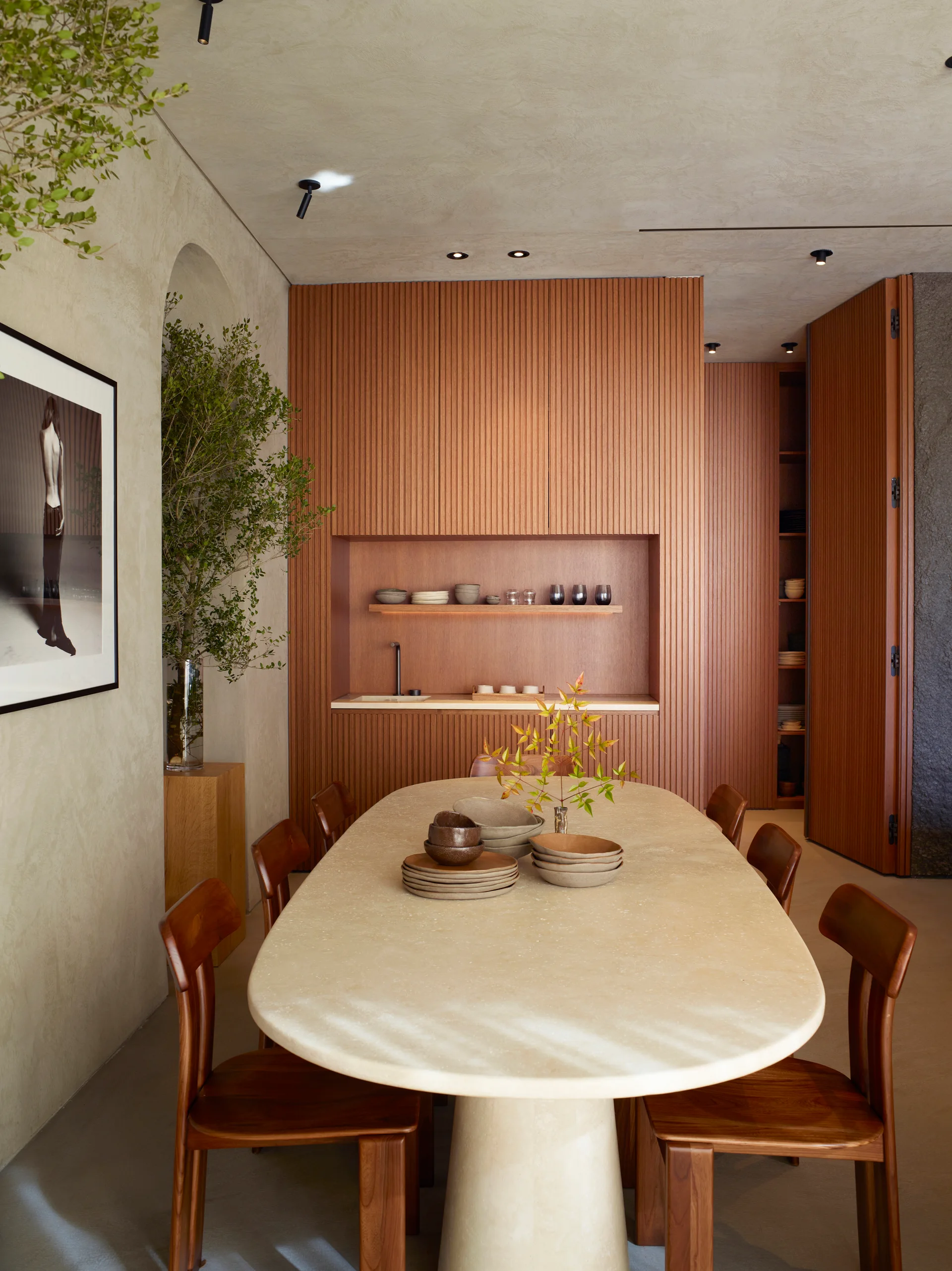 A minimalist dining room features an oval beige stone table, wooden chairs, textured plaster walls, and a built-in kitchenette wall with fluted wood paneling.