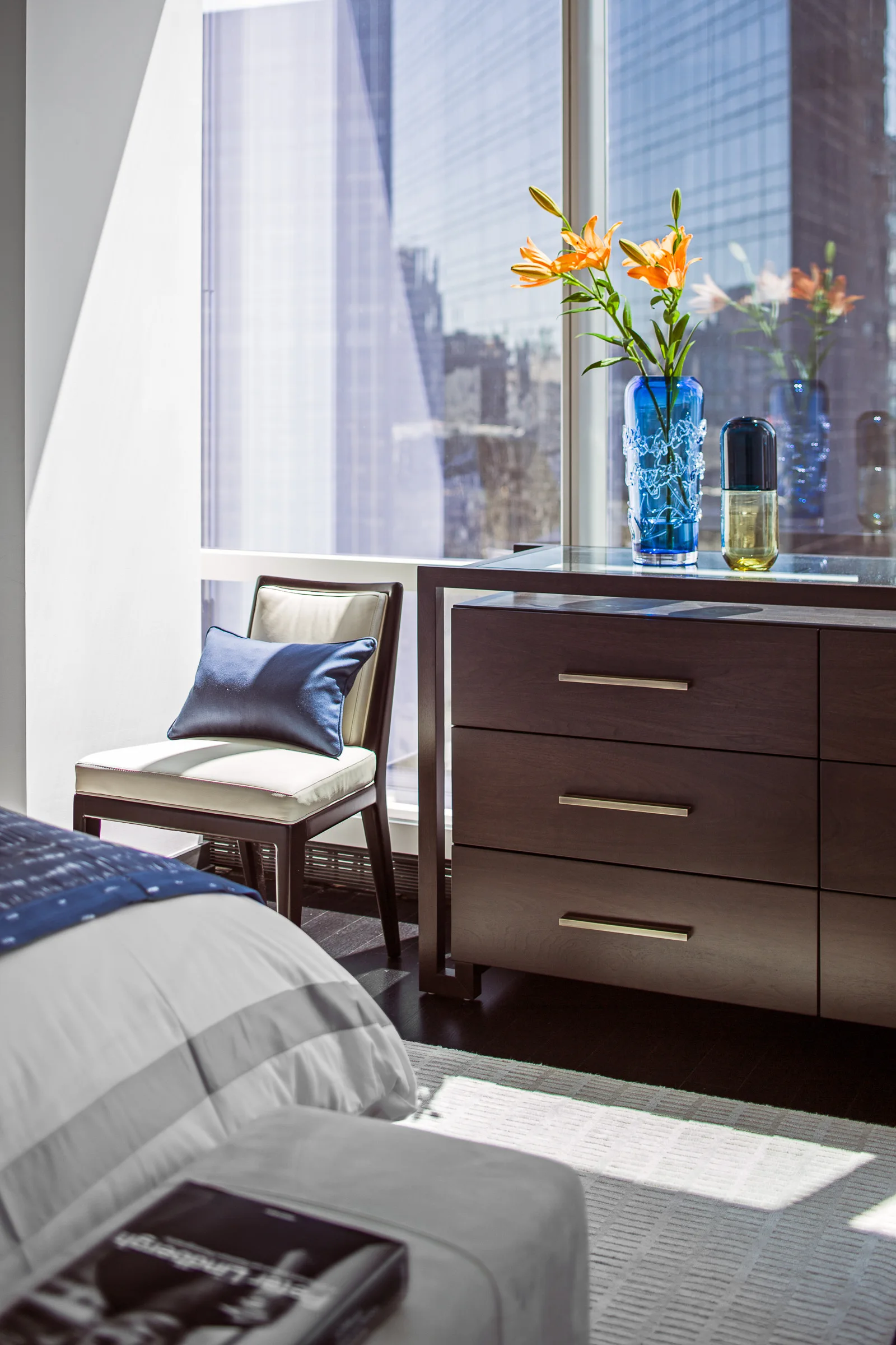 A modern bedroom corner features a dark wood dresser next to an upholstered armchair with a blue pillow in front of a large city window.