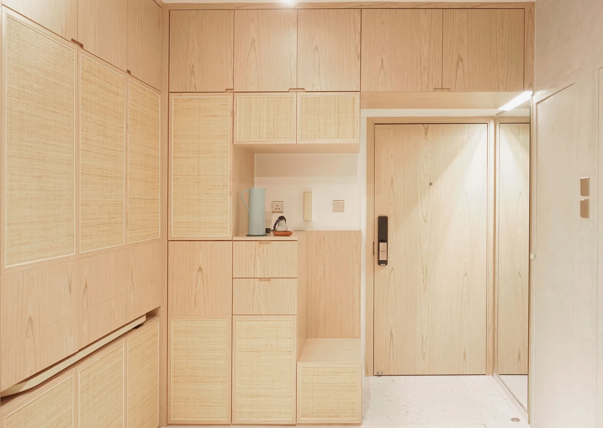 A minimalist entryway features custom light wood built-in cabinets with woven rattan panels, an integrated bench, and a tall mirror.