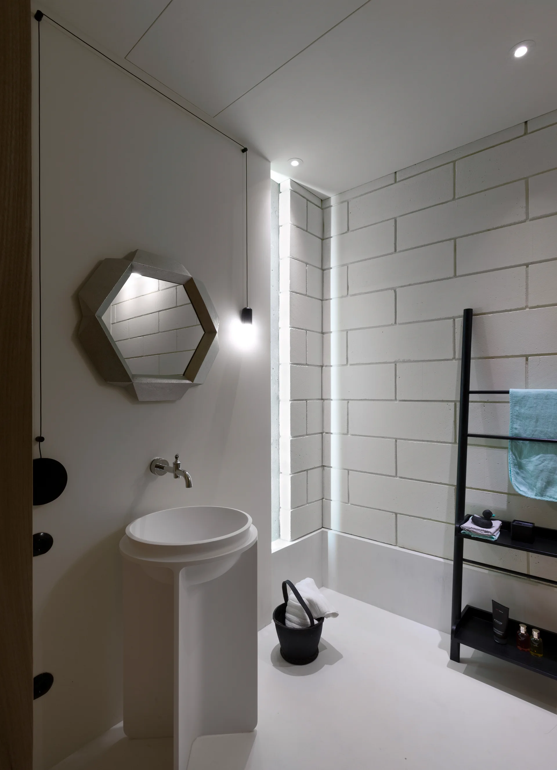 A minimalist white bathroom features a sculptural pedestal sink under a geometric mirror, a painted cinder block wall, and a black ladder towel rack.