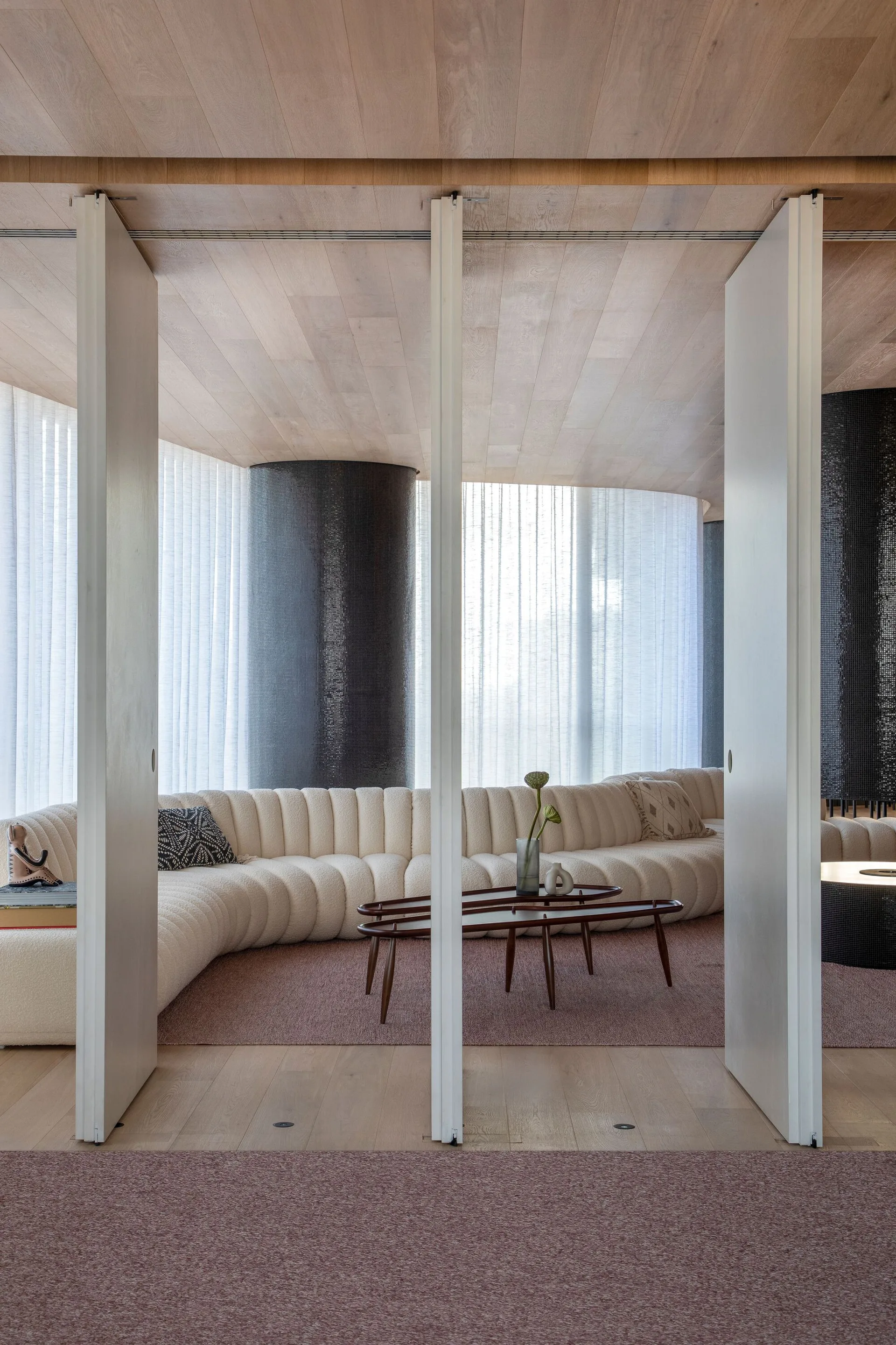 A modern living area featuring a curved cream bouclé sofa, low nesting coffee tables, and dark mosaic columns behind sheer white curtains.