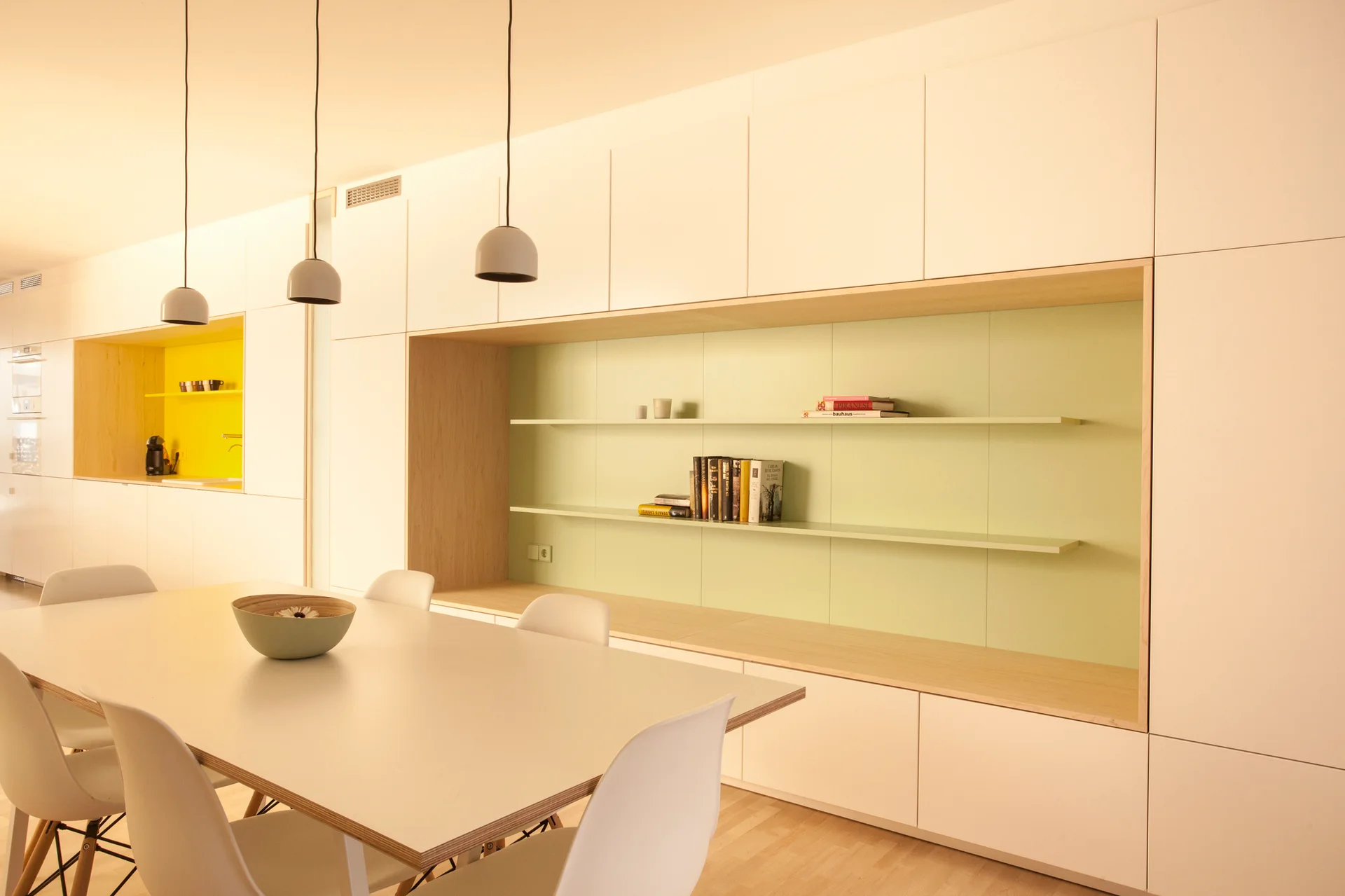 A minimalist dining room with a white dining table, modern chairs, and a large built-in storage wall featuring a recessed pale green display niche.