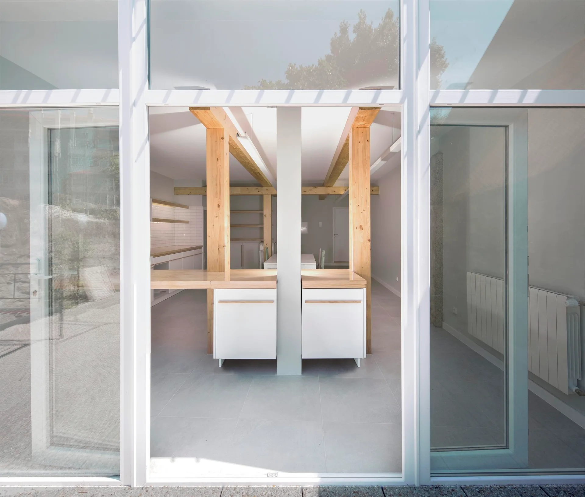 A modern minimalist kitchen with white cabinets, wood countertops, and exposed wooden beams is seen through a large glass door facade.