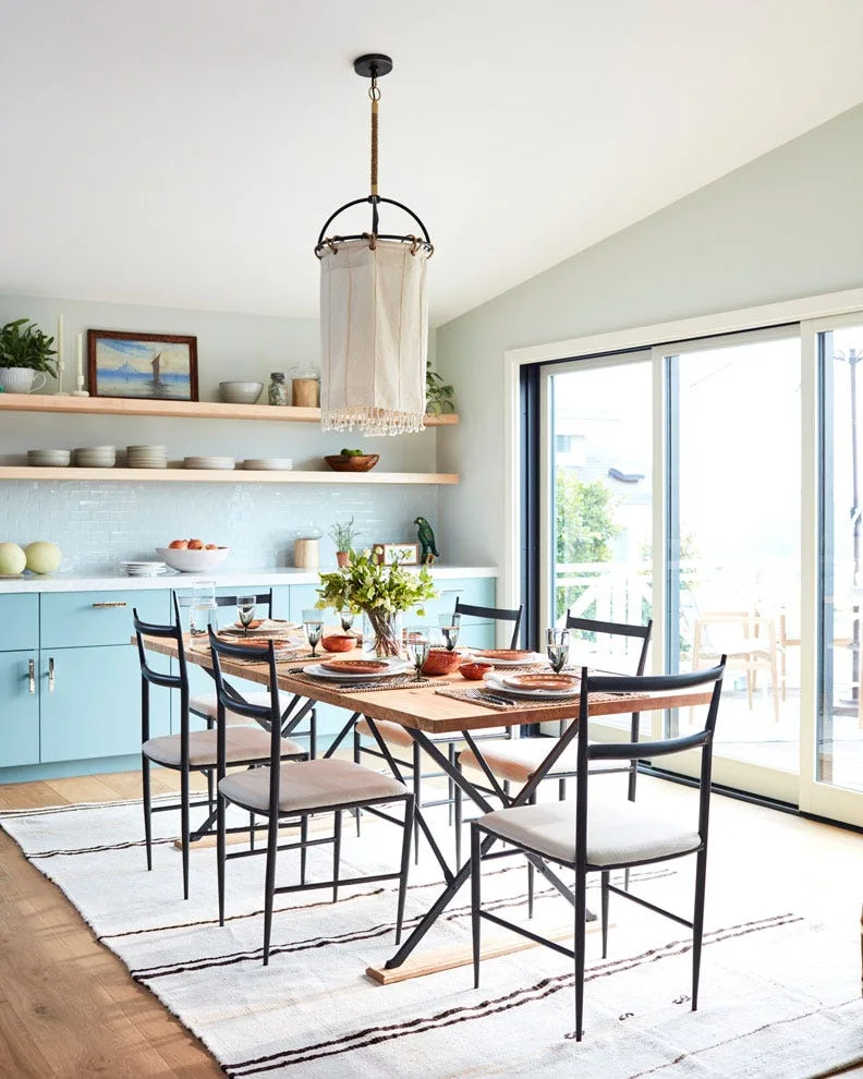 A dining room features a wooden table with black chairs situated next to a light blue kitchen counter and sliding glass doors.