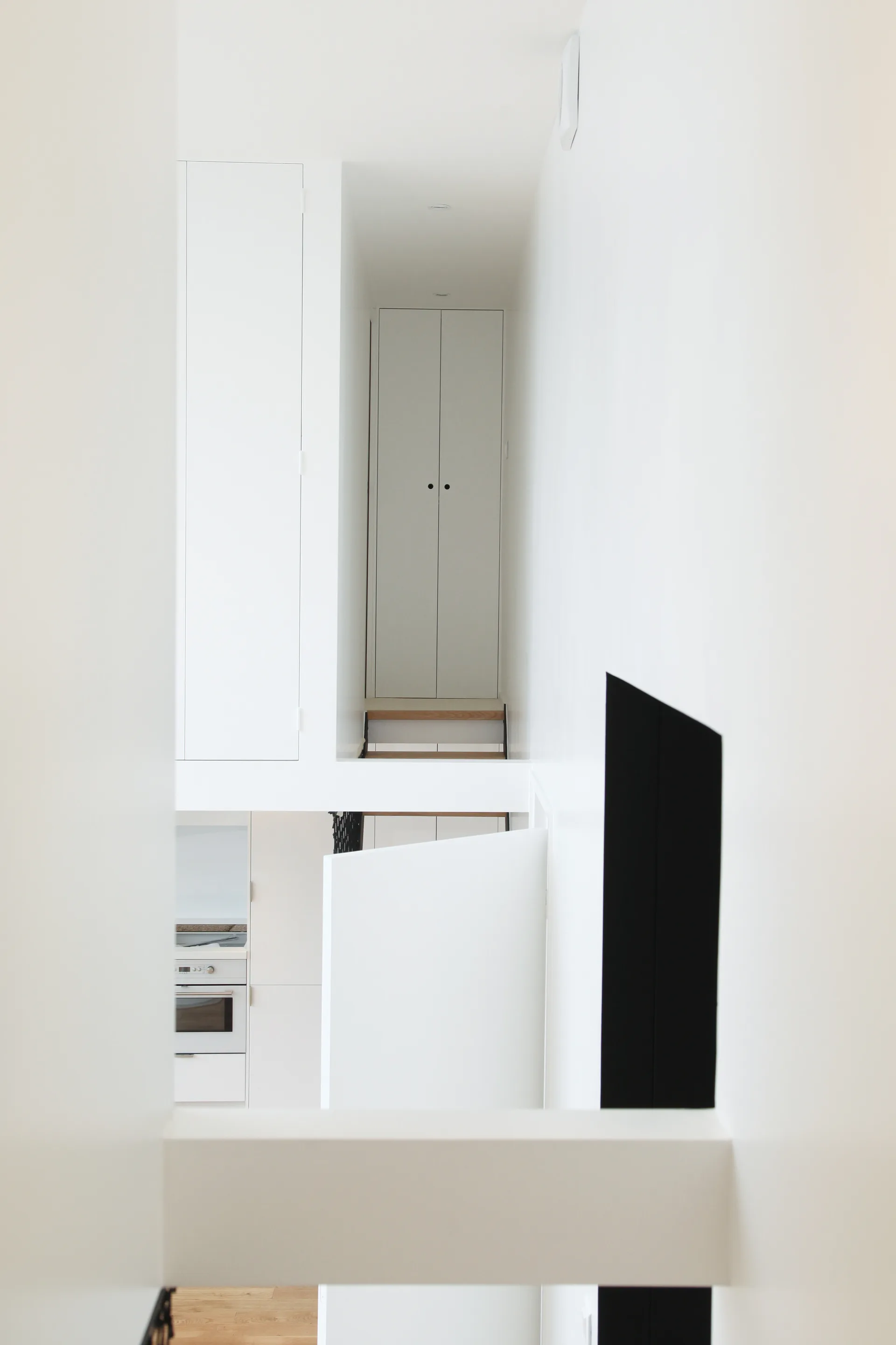 A minimalist white split-level hallway features built-in flat-panel cabinets, integrated wood stairs, and a glimpse of a kitchen below.