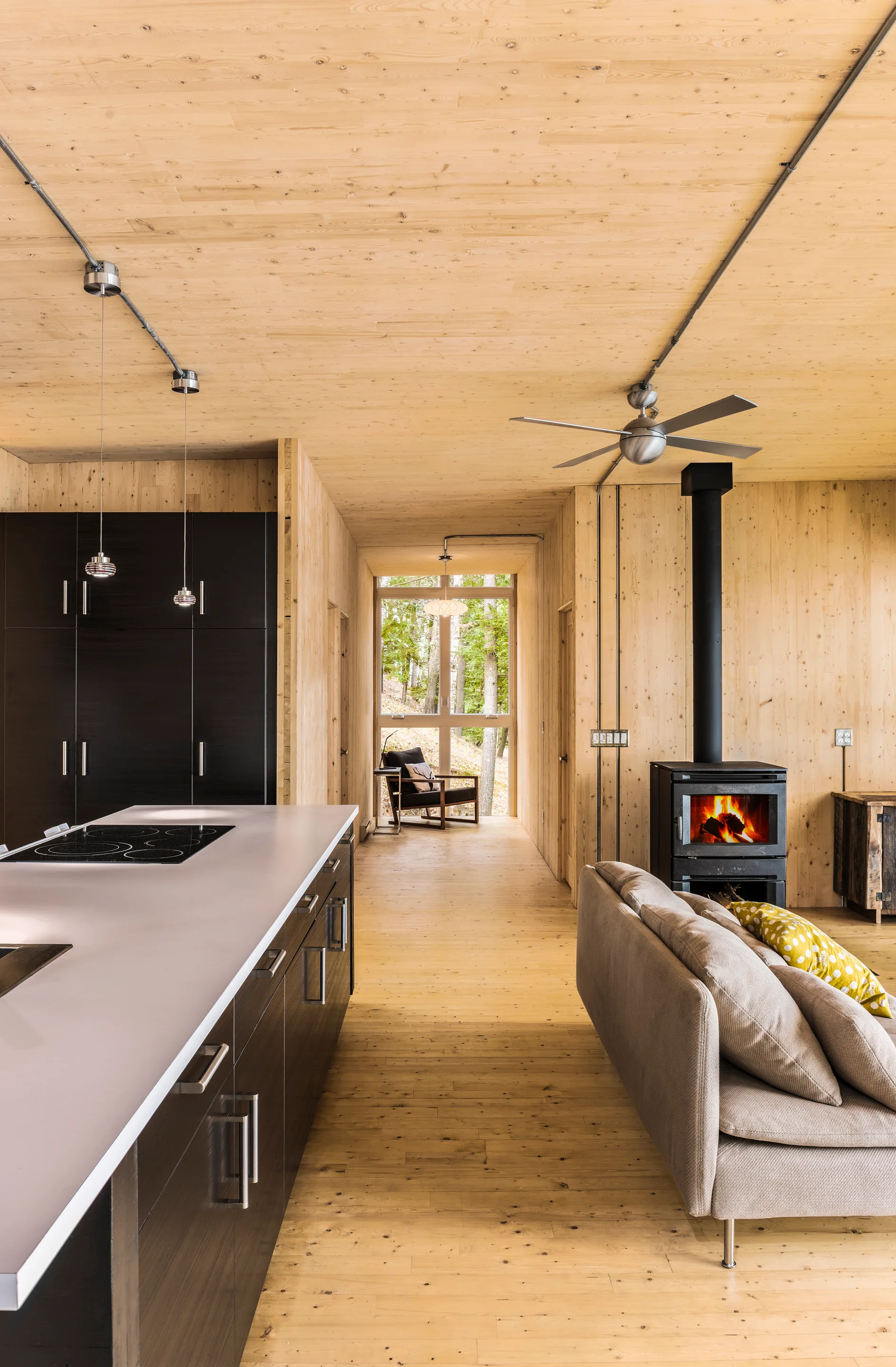 An open-concept kitchen and living area features light wood paneling on the walls and ceiling, a dark kitchen island, and a black wood-burning stove.