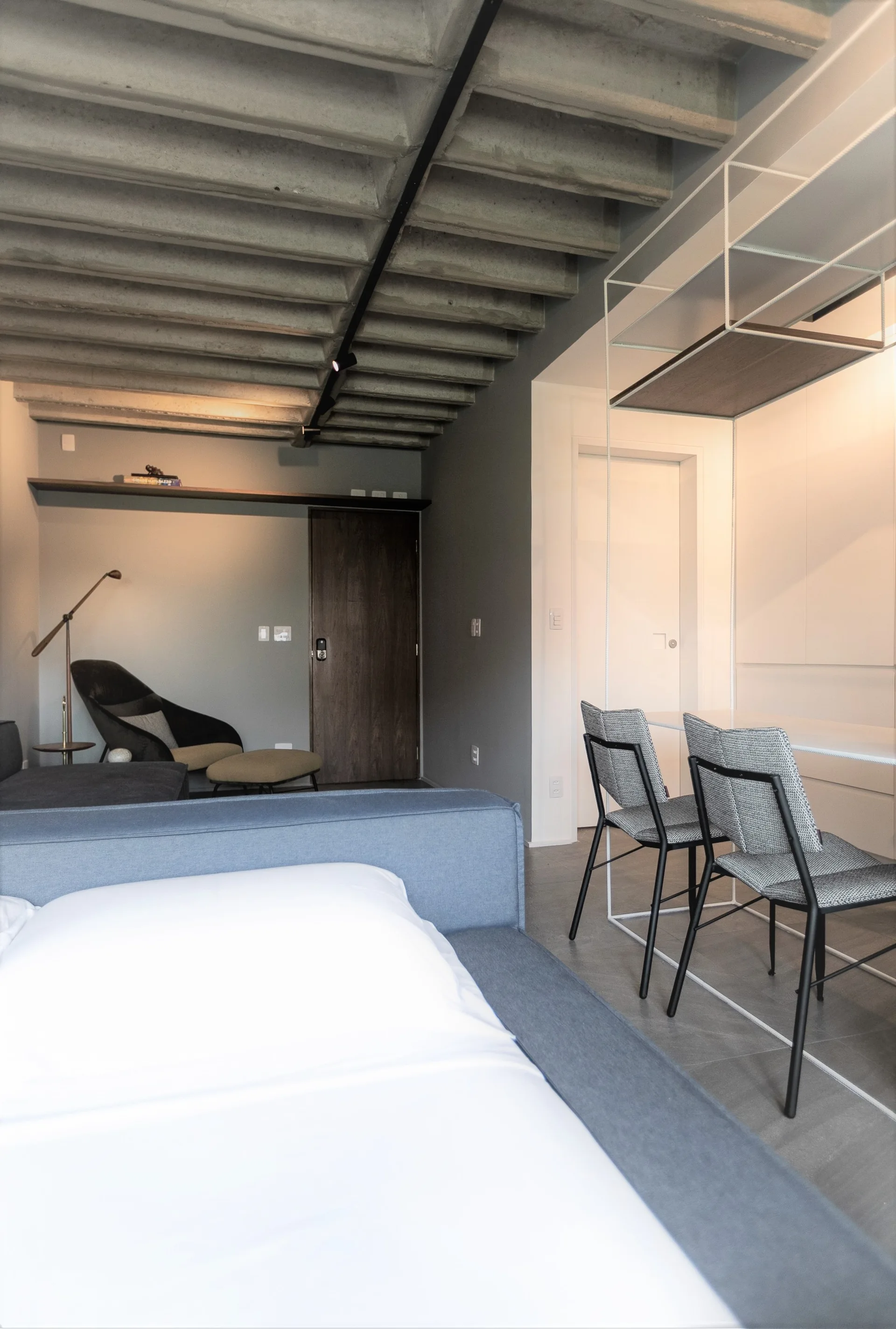 A minimalist bedroom featuring an exposed concrete rib ceiling, a blue upholstered bed, and a white metal-framed desk with gray chairs.