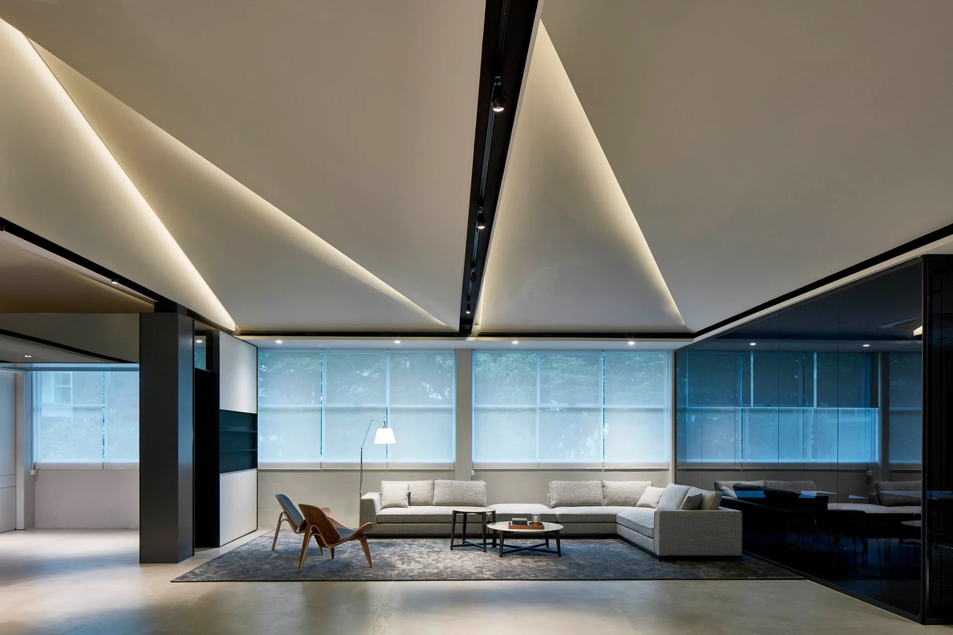A minimalist lounge features a grey sectional sofa and wooden chairs under an angled geometric ceiling with built-in cove lighting.