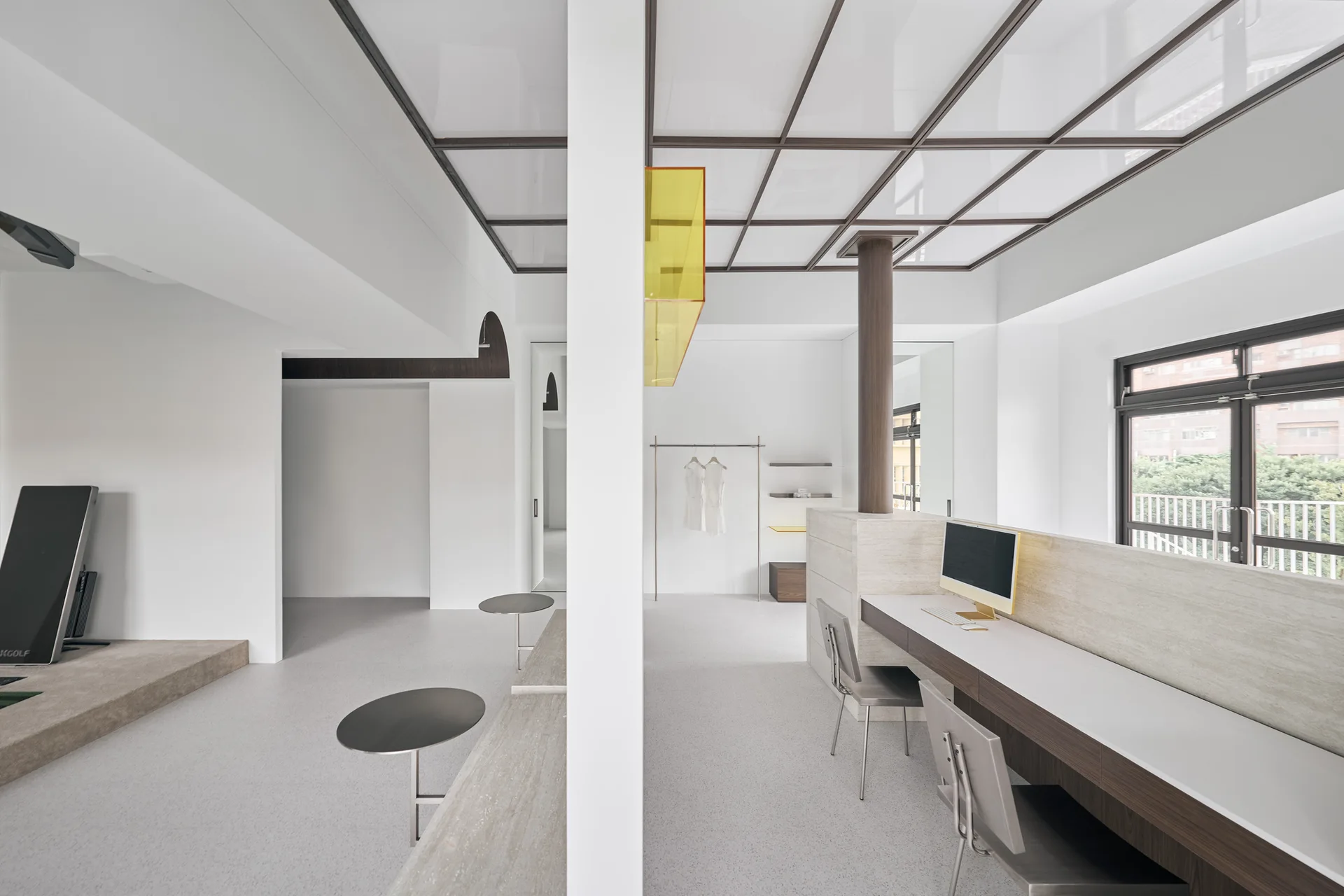 A minimalist white office space features a long stone-like built-in desk, two metal chairs, a central white partition, and a dark geometric ceiling grid.